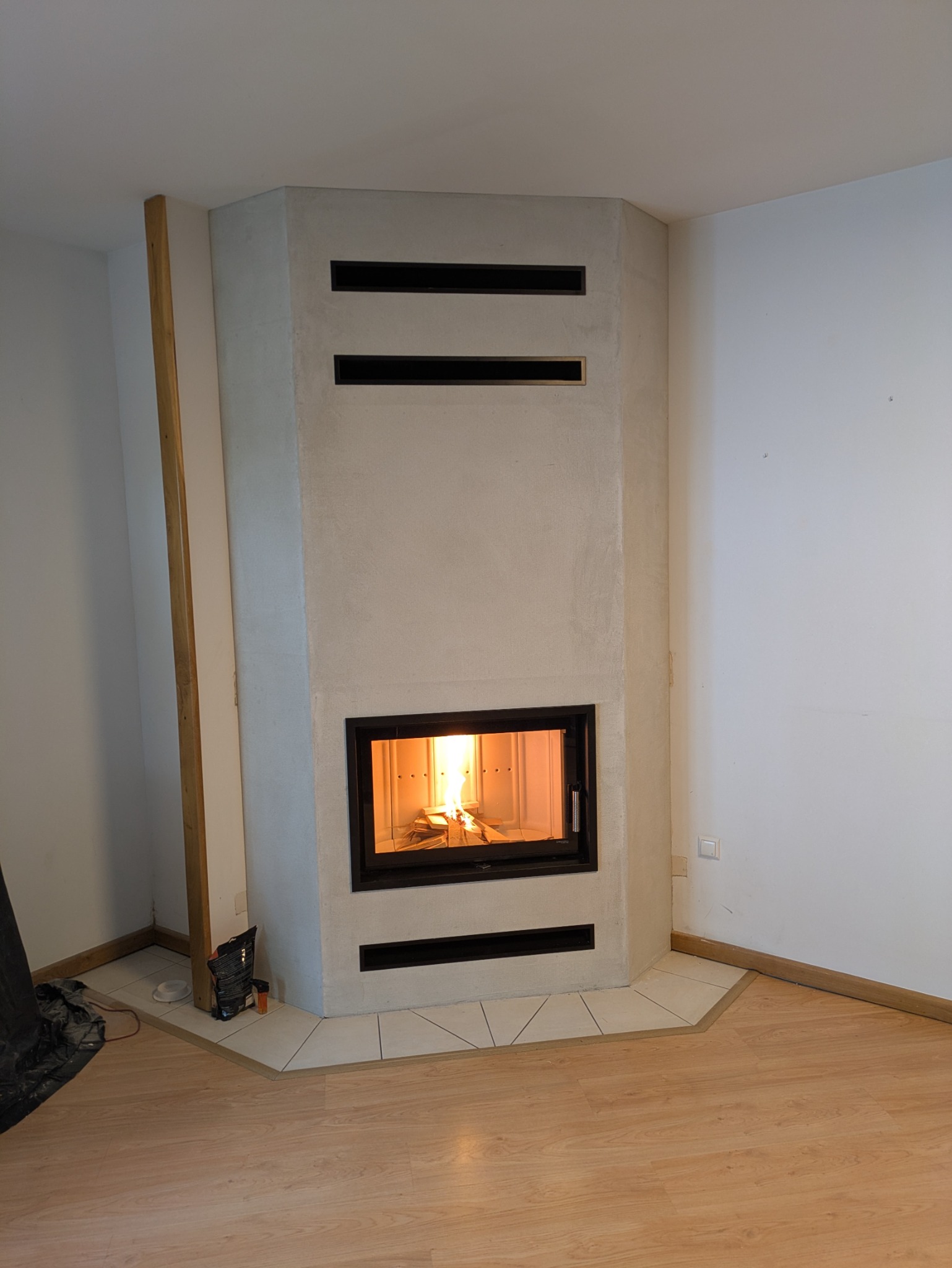 Narożny kominek z paleniskiem w obudowie z beżowego tynku strukturalnego, z trzema poziomymi wlotami powietrza. Podłoga wyłożona panelami i płytkami.