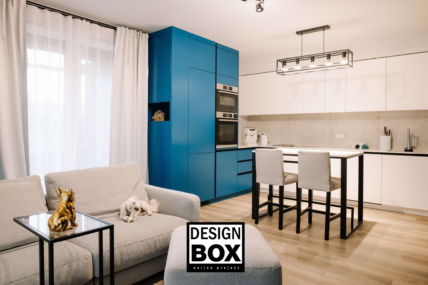 Nowoczesne wnętrze: błękitne szafki kuchenne, białe fronty, wyspa z krzesłami, sofa z psem i złotą figurką, jasna podłoga, minimalistyczne oświetlenie.