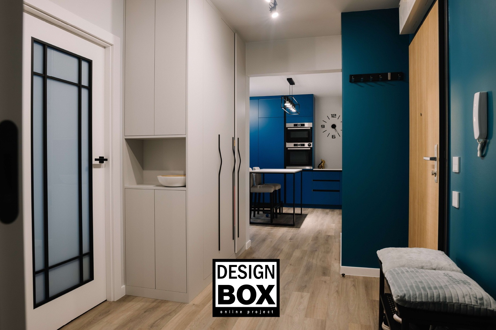 Nowoczesny przedpokój z zabudową w odcieniach szarości, akcentem niebieskiej kuchni w tle i drewnianymi drzwiami wejściowymi. Minimalistyczny design.