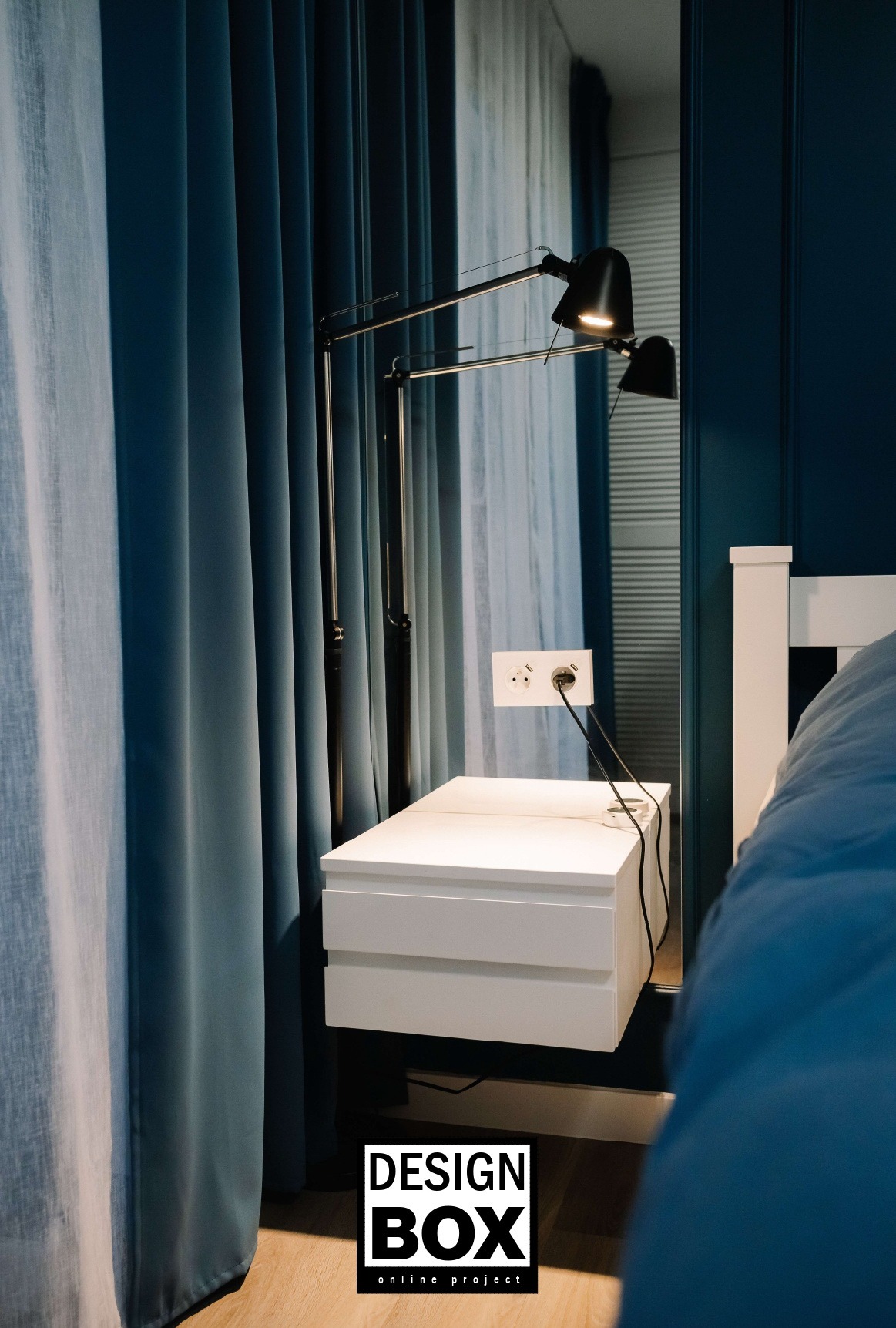 Stylowa sypialnia: biała szafka nocna z lampką i gniazdkiem na tle błękitnych zasłon i granatowej ściany. Minimalistyczny design z logo Design Box.