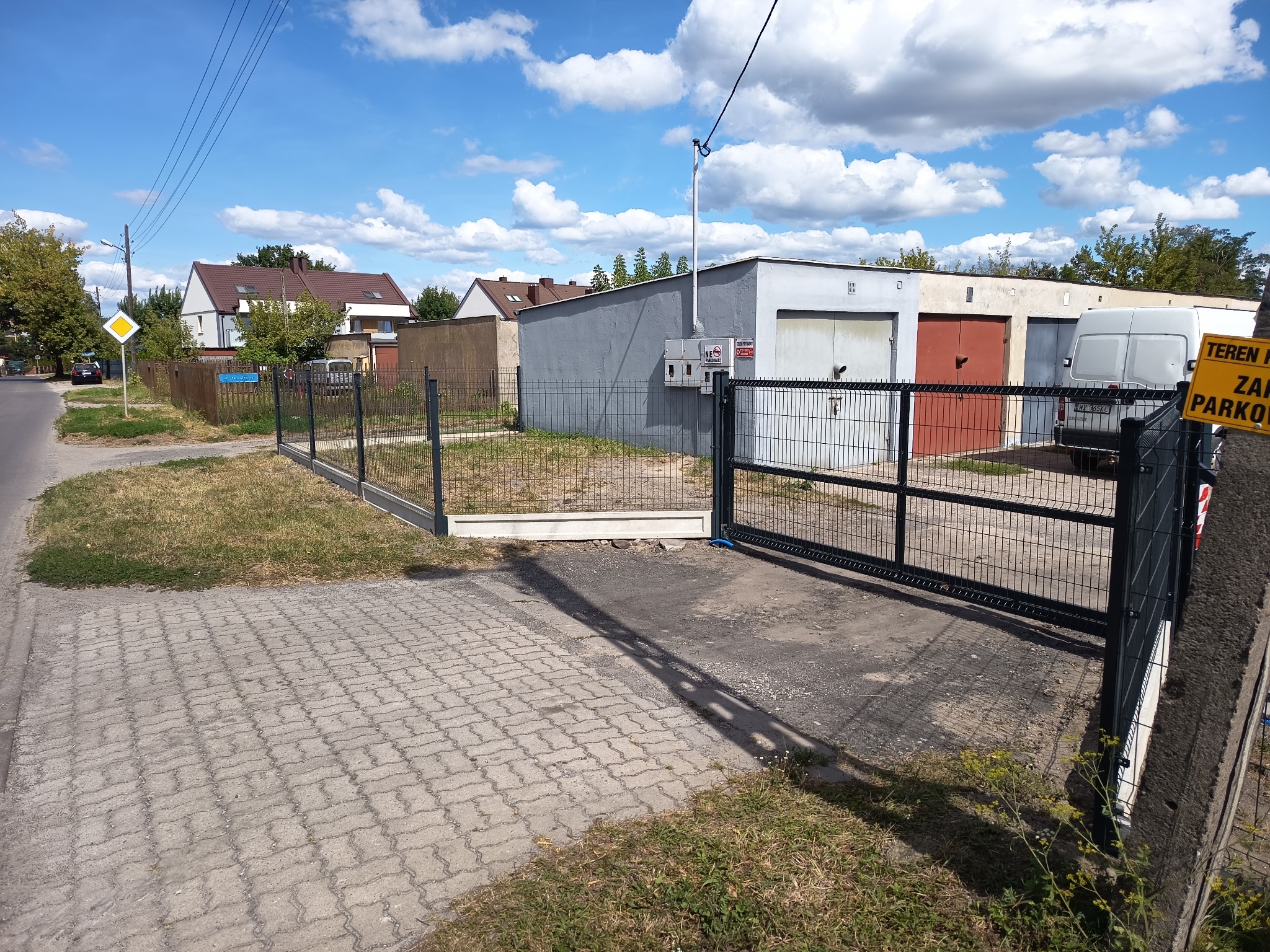 Nowoczesne, metalowe ogrodzenie panelowe z bramą wjazdową na posesję. Widoczne budynki garażowe w tle i błękitne niebo z chmurami.