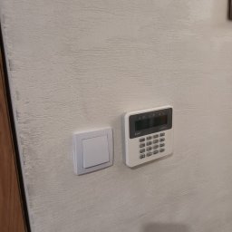 OPSERWIS - Panel alarmowy z klawiaturą numeryczną marki Safer Perfecta, zamontowany na ścianie obok włącznika światła. Teksturowana powierzchnia ściany. Minimalistyczny design.