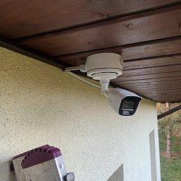 OPSERWIS - Biała kamera monitoringu HiLook zamontowana pod drewnianym okapem budynku. Widoczne kable i fragment drabiny oparty o ścianę.