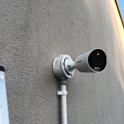 OPSERWIS - Kamera monitoringu Hikvision zamontowana na szarej, chropowatej ścianie budynku. Widoczne elementy montażowe i fragment drabiny po lewej stronie kadru.