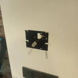 Czarna puszka instalacyjna z kablem sieciowym RJ45 wystającym ze ściany, obok włącznik światła. Montaż gniazdka sieciowego.