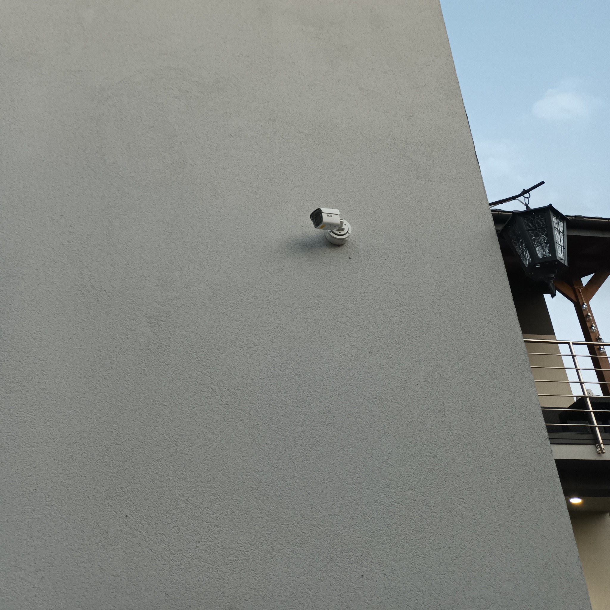Kamera monitoringu zamontowana na szarej elewacji budynku. Widoczna lampa balkonowa w tle. Ujęcie z dołu podkreśla dyskretny montaż.