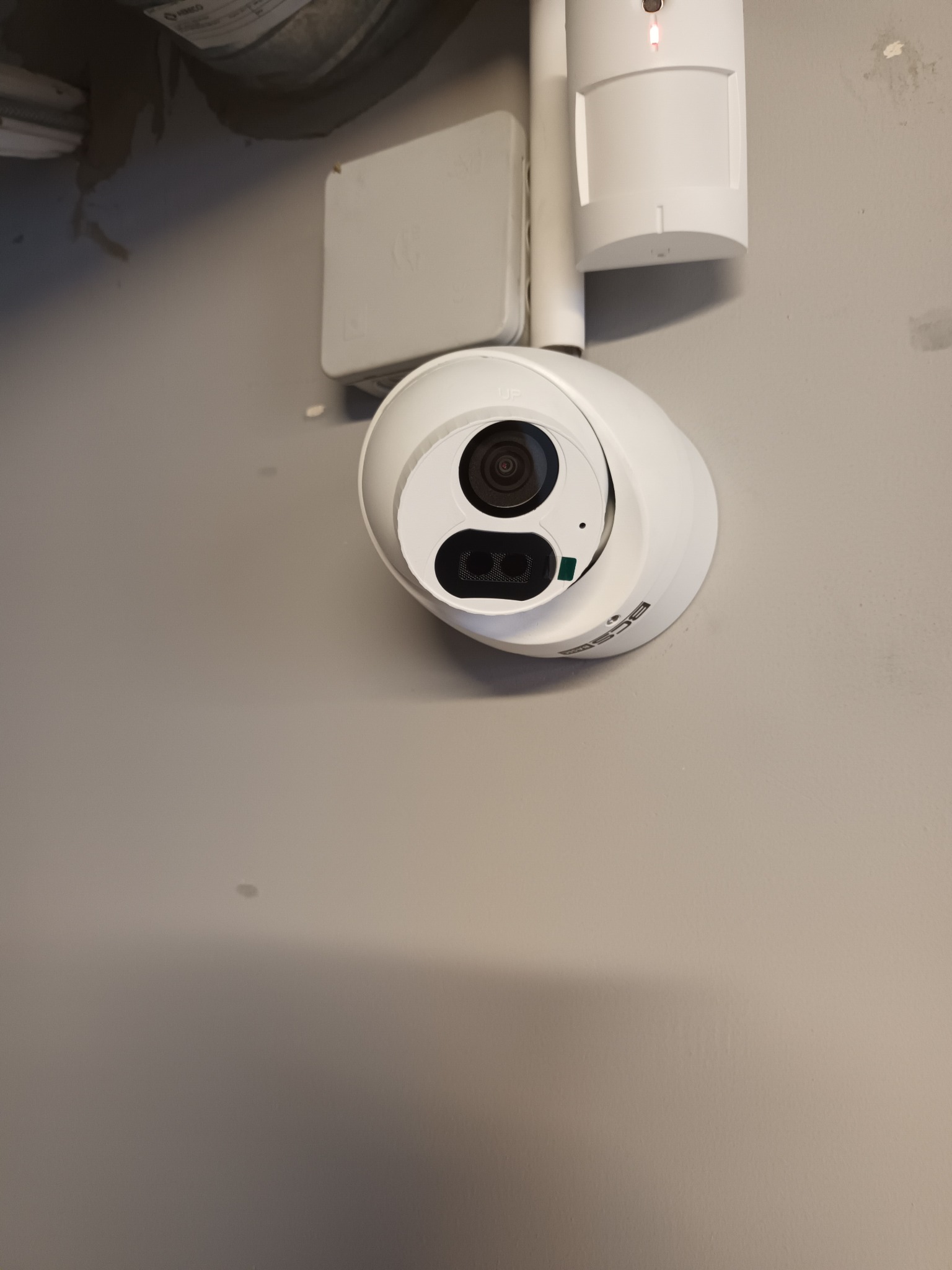 Zamontowana kamera monitoringu BCS na ścianie, obok czujnik ruchu z czerwoną diodą. Widoczne przewody i elementy instalacji. Niewielkie zabrudzenia na ścianie.