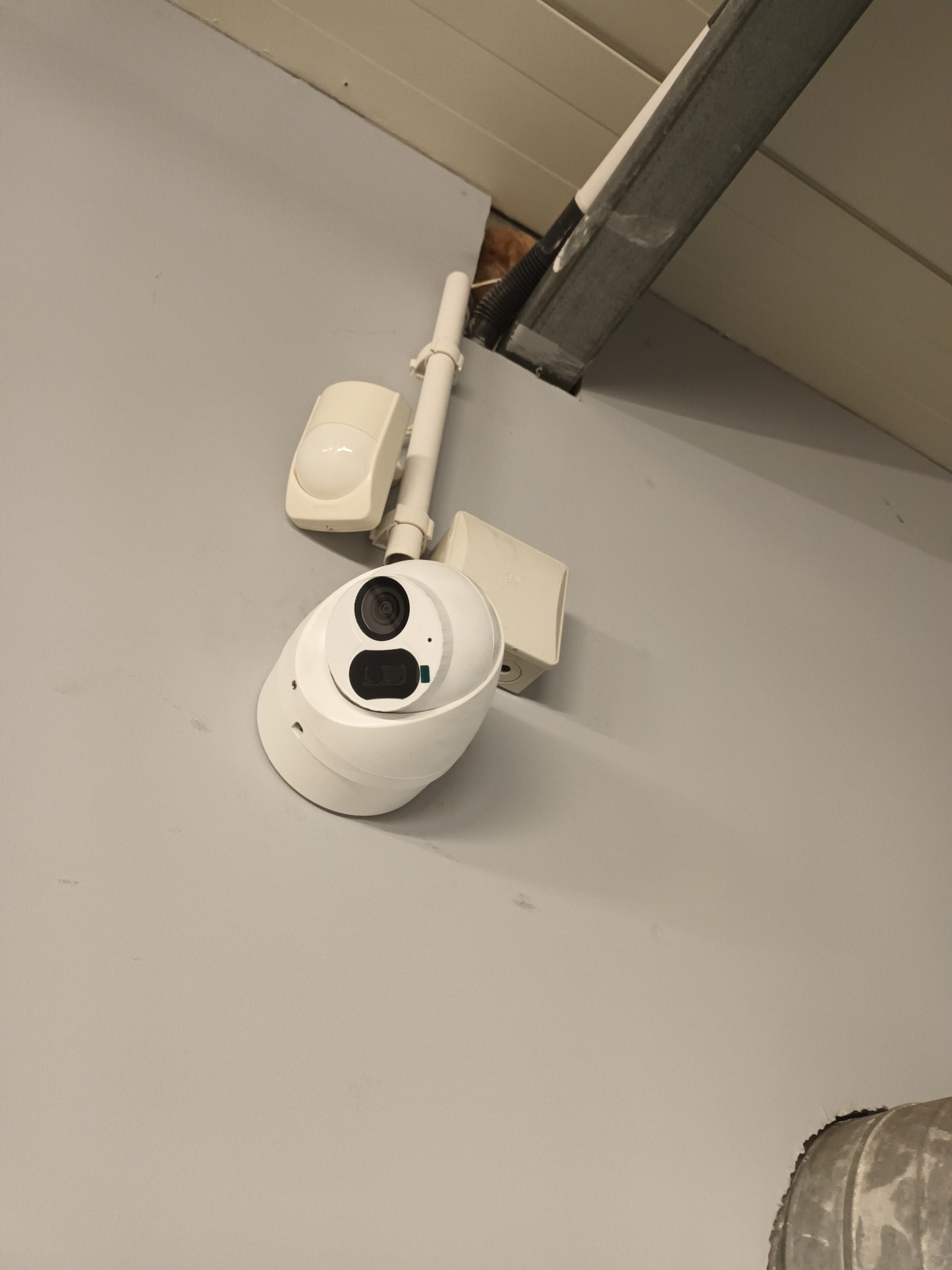 Zamontowana biała kamera monitoringu z czujnikiem ruchu na szarej ścianie, widoczne kable i fragment sufitu. Ujęcie z dołu.