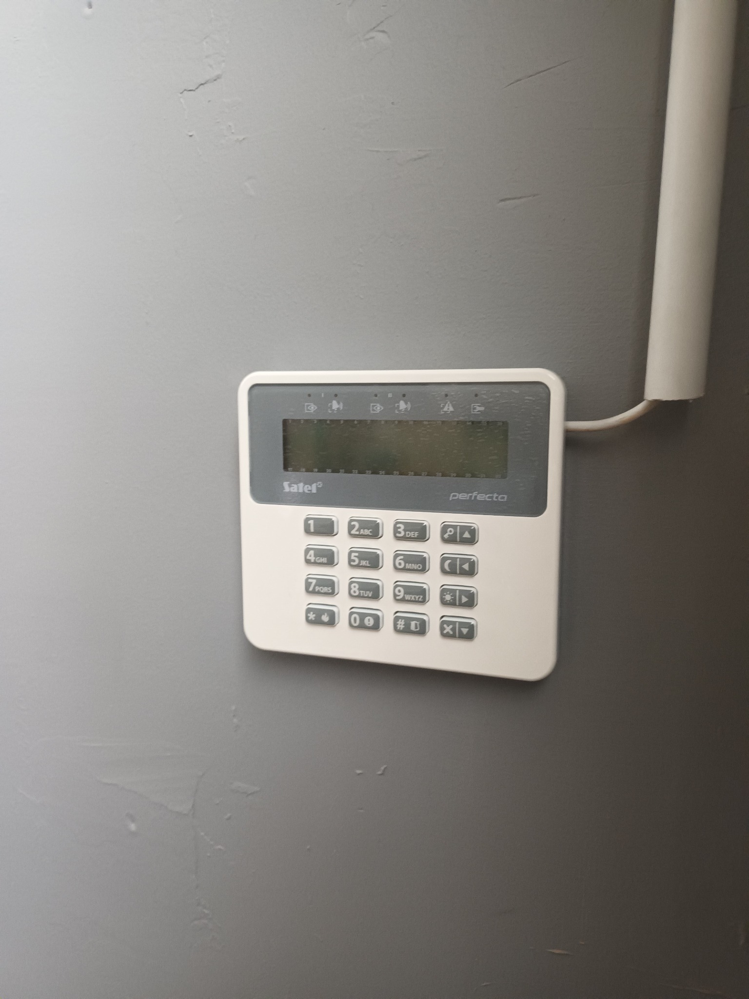 Panel sterowania alarmem Satel Perfecta, zamontowany na szarej ścianie, z widocznym przewodem. Klawiatura numeryczna i wyświetlacz.