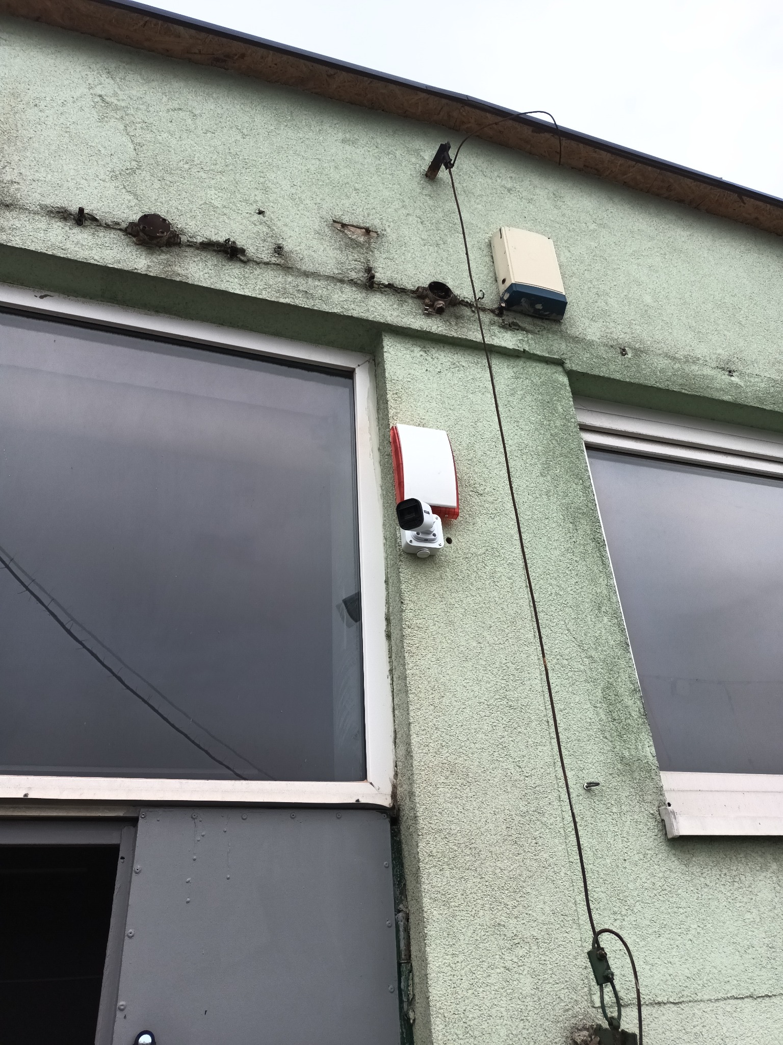 Kamera monitoringu i sygnalizator alarmowy na elewacji budynku z oknami. Widoczne ślady po wcześniejszych instalacjach.