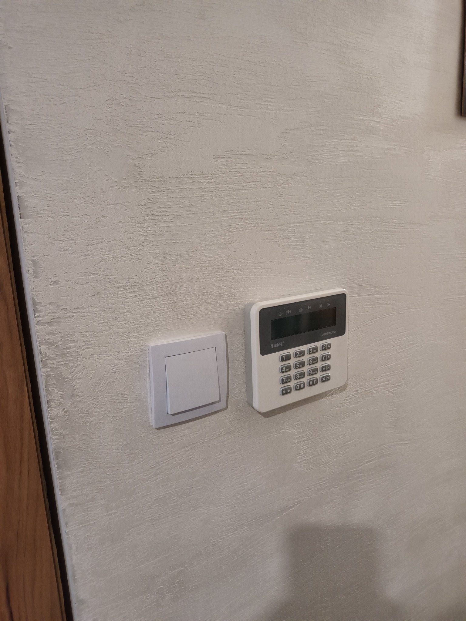 Panel alarmowy z klawiaturą numeryczną marki Safer Perfecta, zamontowany na ścianie obok włącznika światła. Teksturowana powierzchnia ściany. Minimalistyczny design.