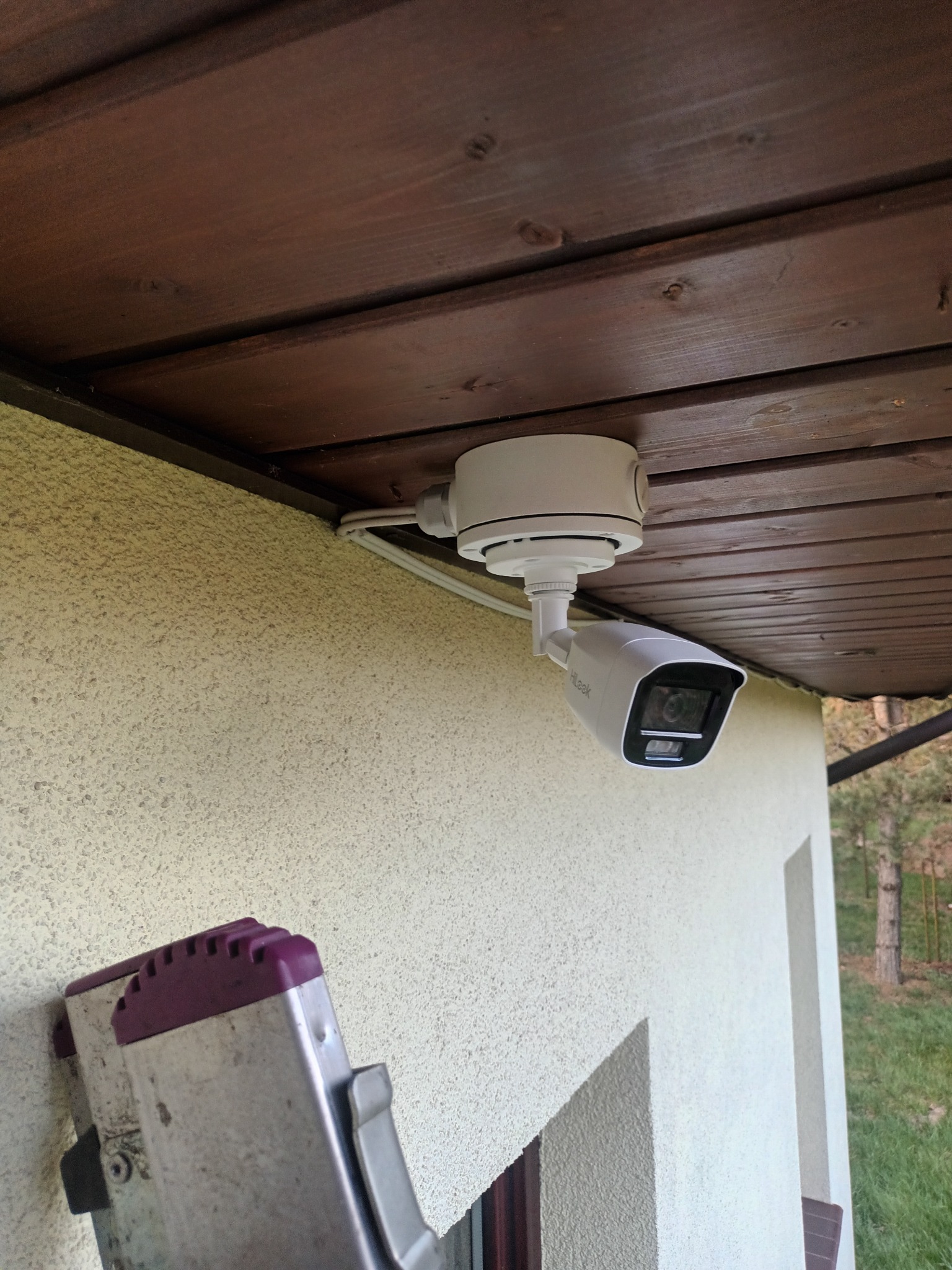 Biała kamera monitoringu HiLook zamontowana pod drewnianym okapem budynku. Widoczne kable i fragment drabiny oparty o ścianę.