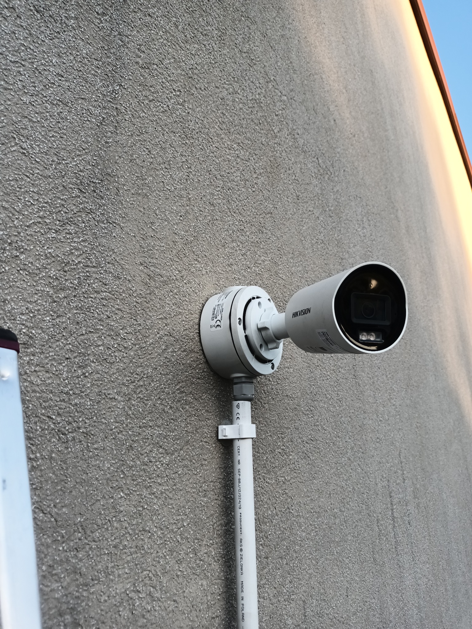 Kamera monitoringu Hikvision zamontowana na szarej, chropowatej ścianie budynku. Widoczne elementy montażowe i fragment drabiny po lewej stronie kadru.
