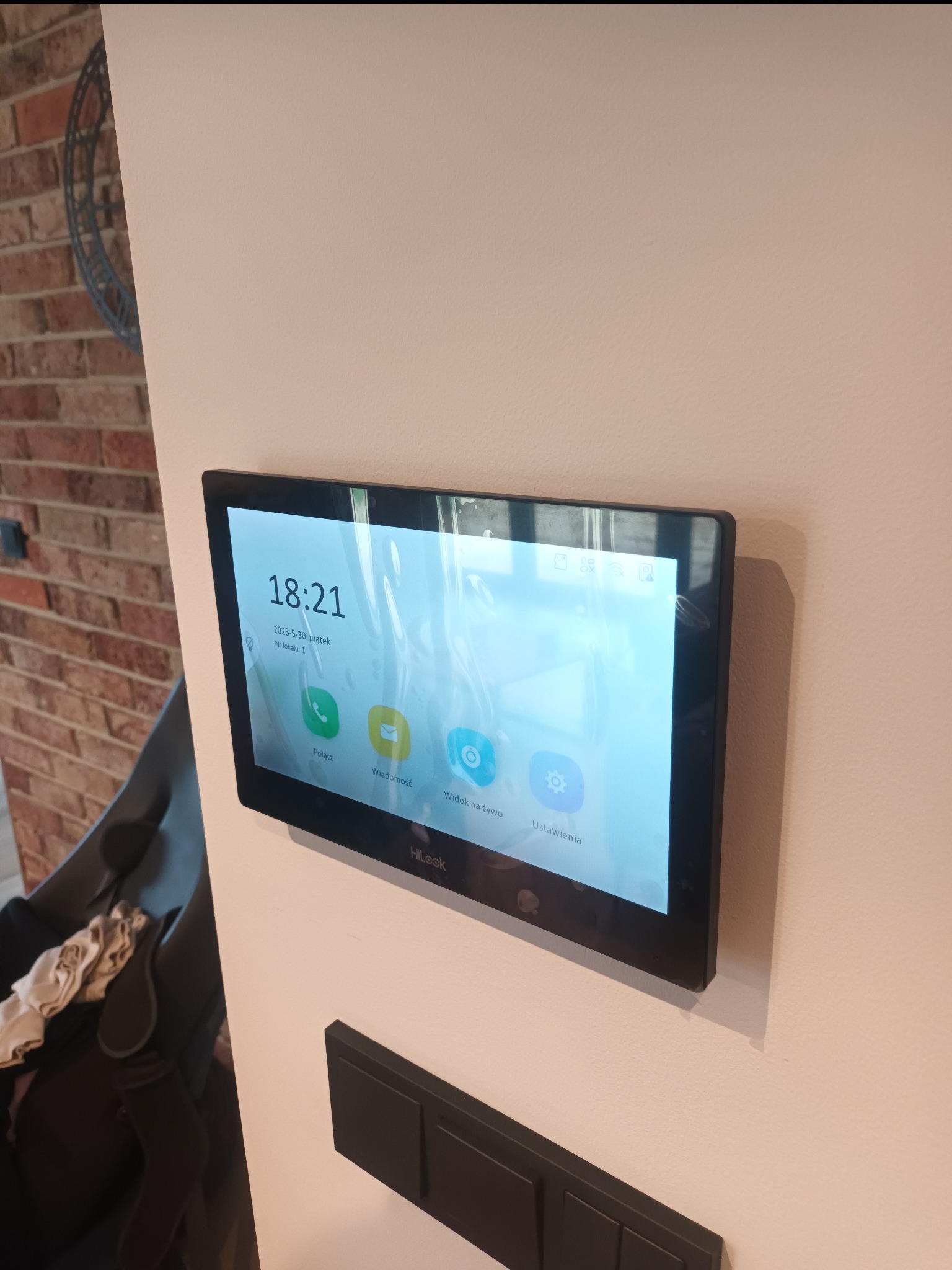 Zamontowany na ścianie panel wideodomofonu HiLook z interfejsem użytkownika pokazującym aktualną godzinę i opcje połączeń, obok włącznik światła.