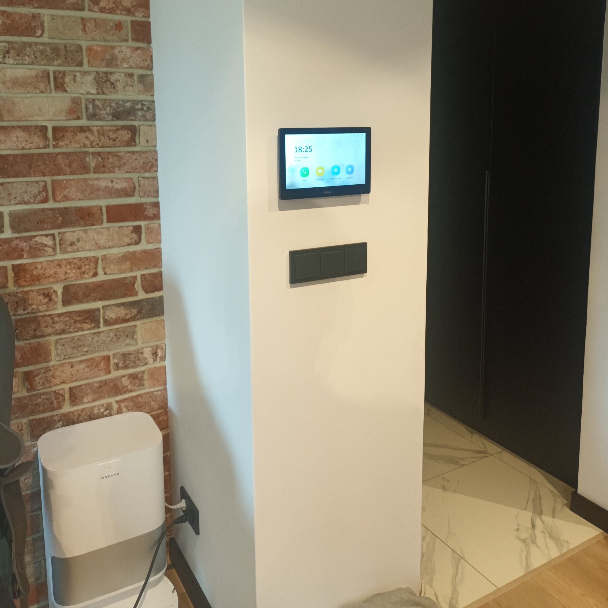 Panel sterowania inteligentnego domu zamontowany na ścianie obok włączników, z widocznym ekranem i ikonami aplikacji. Obok ściana z cegły i robot sprzątający.