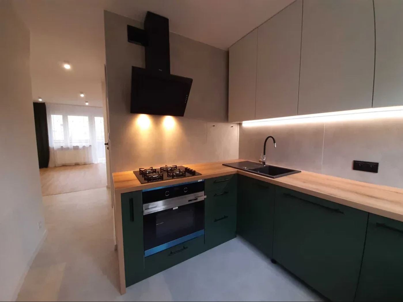 Nowoczesna kuchnia z zielonymi szafkami i drewnianym blatem. Czarny okap nad płytą gazową. W tle widoczny salon z oknami.