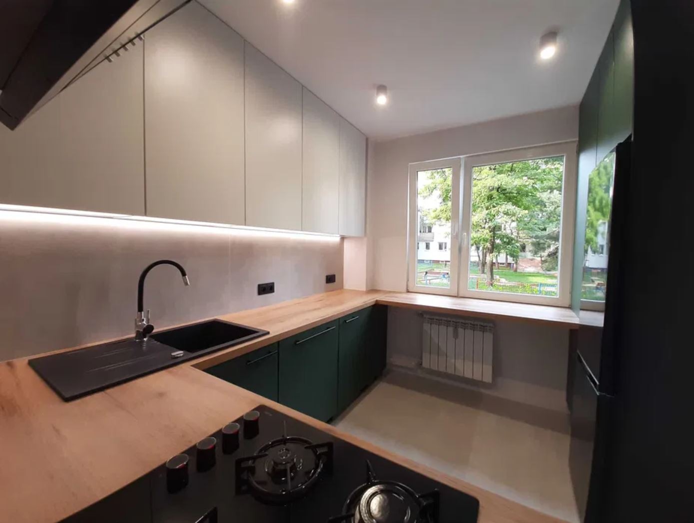 Nowoczesna kuchnia z zielonymi szafkami i drewnianym blatem. Widok na zieleń za oknem. Minimalistyczny design i funkcjonalne rozwiązania.