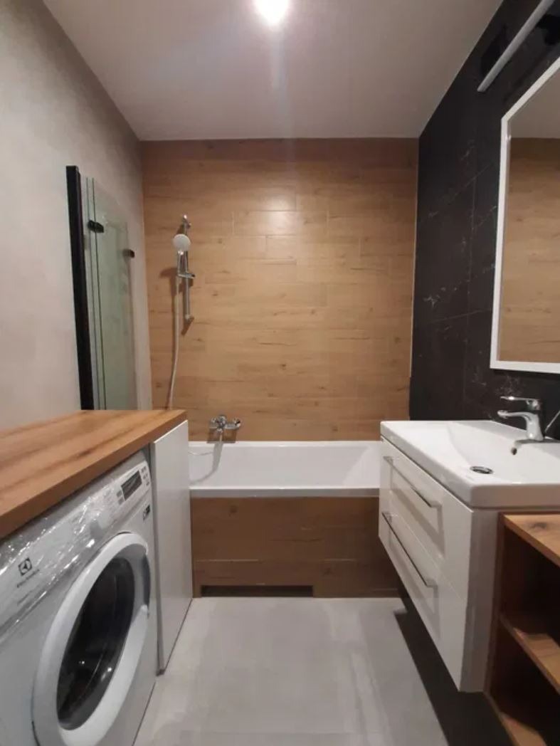 Nowoczesna łazienka z pralką pod blatem, wanną obudowaną drewnem i umywalką z szafką na tle drewnopodobnych płytek. Czarna ściana z lustrem.