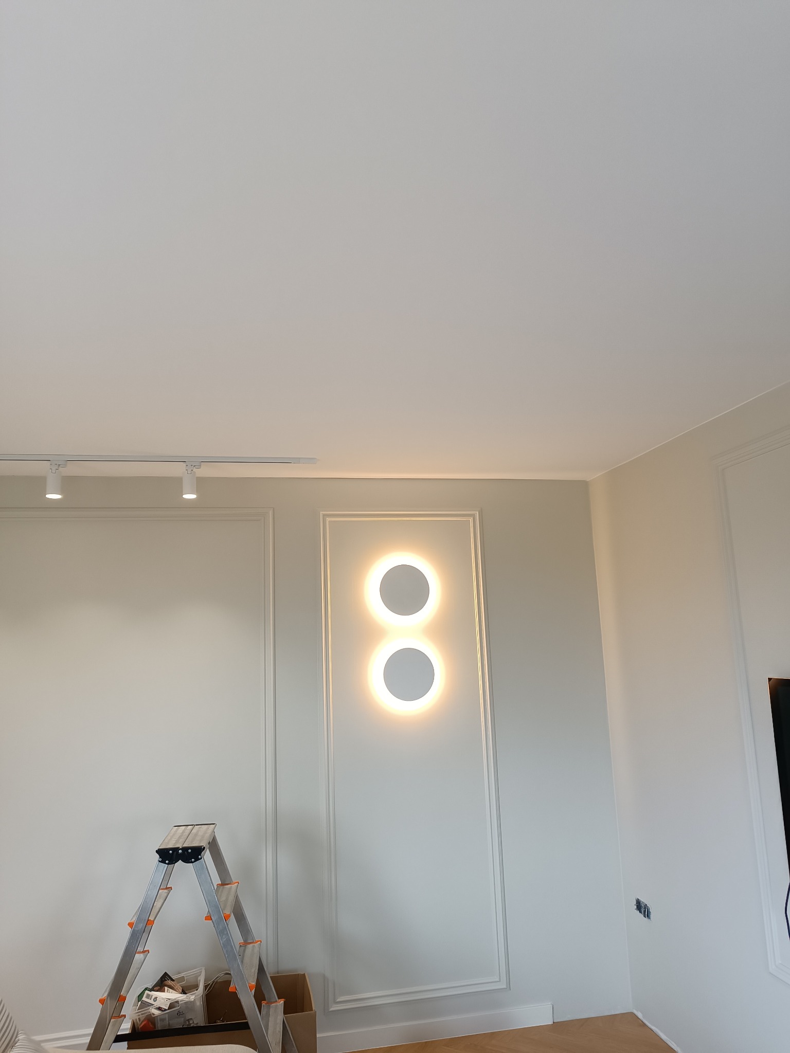 Wnętrze po montażu oświetlenia: dwa okrągłe, podświetlane panele na ścianie z dekoracyjną sztukaterią. Widoczna drabina i listwa sufitowa z reflektorami.