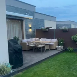 Nowoczesny taras z deską kompozytową: meble ogrodowe z rattanu, kwiaty w donicach na brązowym panelu. Grill z pokrowcem po lewej. Trawnik. Wieczorny kadr.