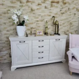 Biała komoda z dekoracjami (figurki, kwiaty) na tle kamiennej ściany i różowego fotela. Mebel z frezowanymi drzwiami i metalowymi uchwytami.