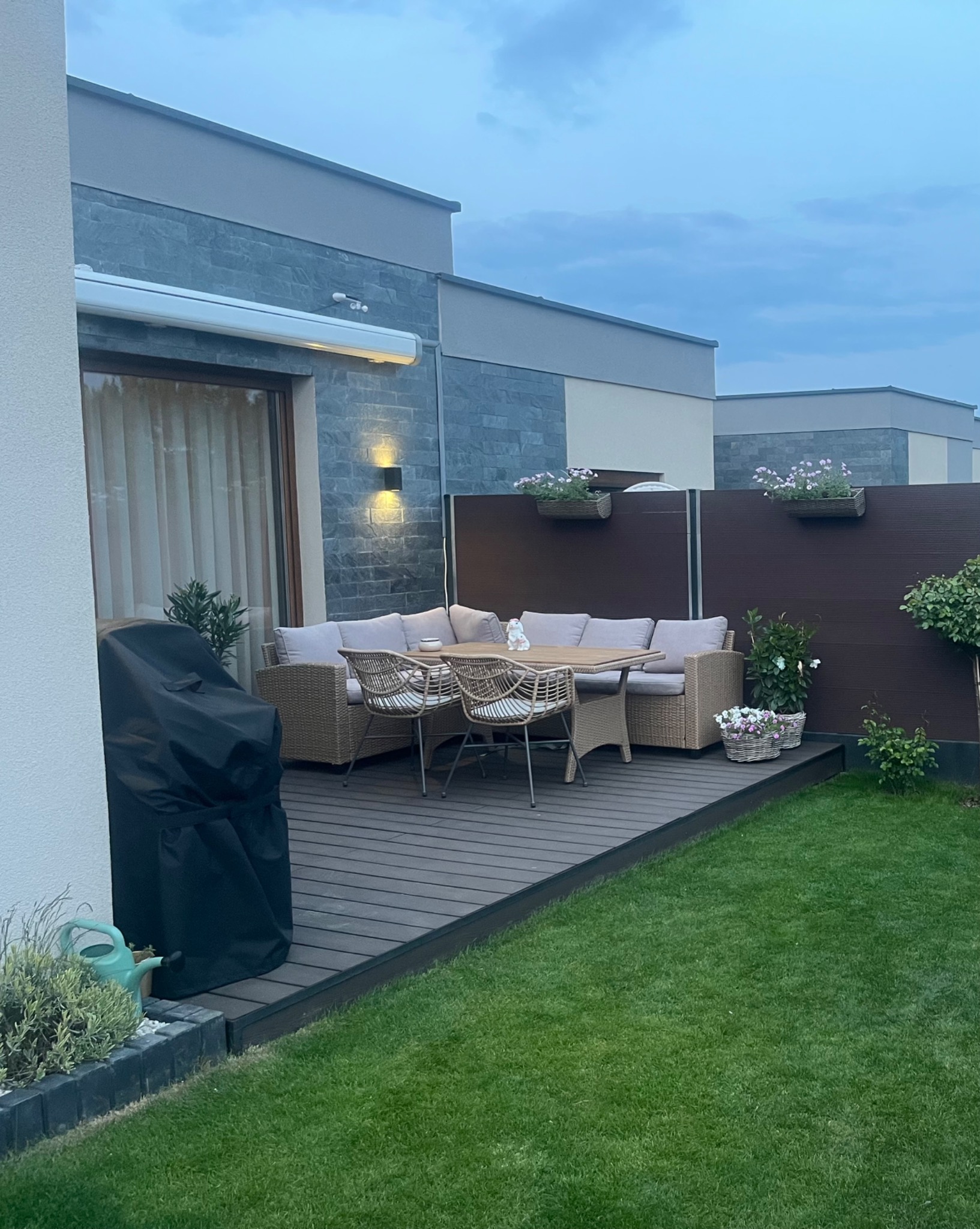 Nowoczesny taras z deską kompozytową: meble ogrodowe z rattanu, kwiaty w donicach na brązowym panelu. Grill z pokrowcem po lewej. Trawnik. Wieczorny kadr.
