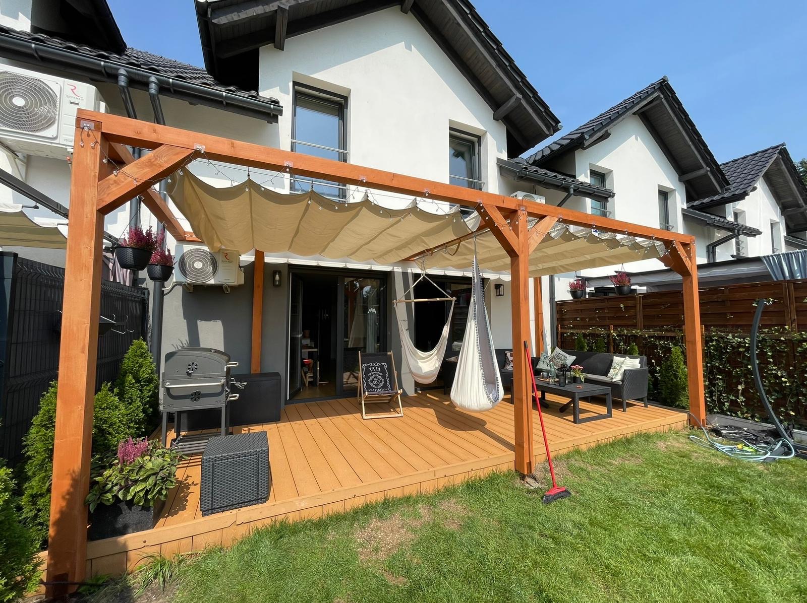 Drewniana pergola z beżowym zadaszeniem nad tarasem, hamaki wiszące, grill, meble ogrodowe, trawnik, dom w tle. Słoneczny dzień.