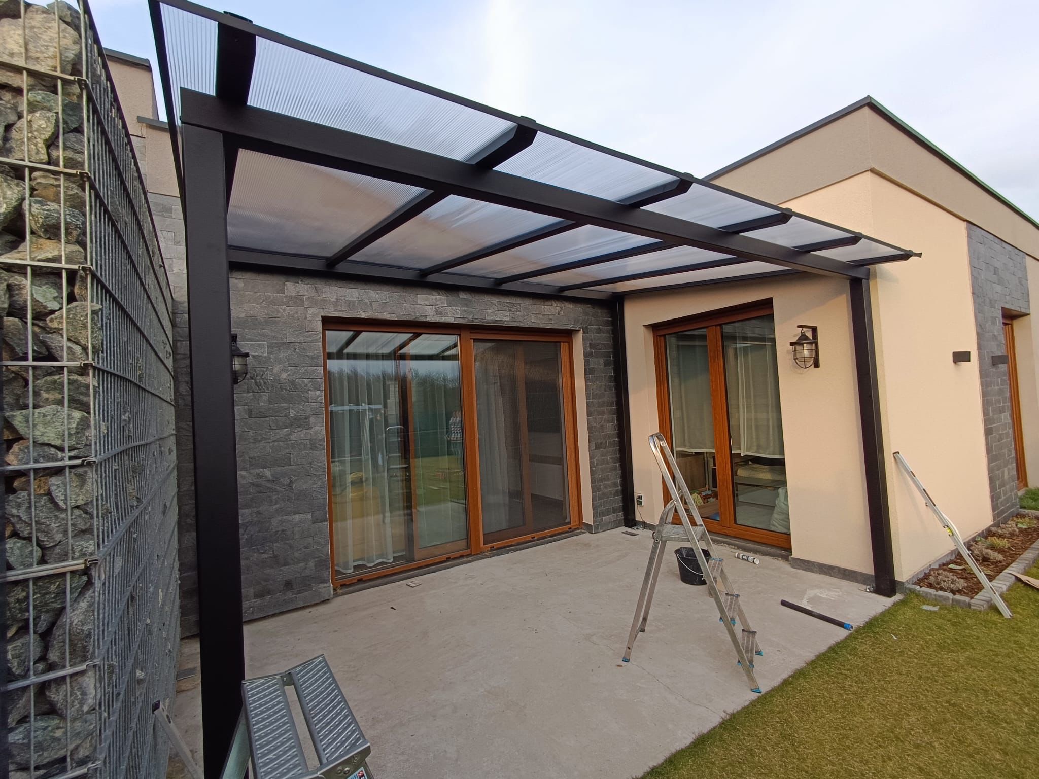 Nowoczesna, czarna pergola z przezroczystym zadaszeniem, przylegająca do budynku z kamienną ścianą i gabionem. Widoczne drabiny i elementy montażowe.