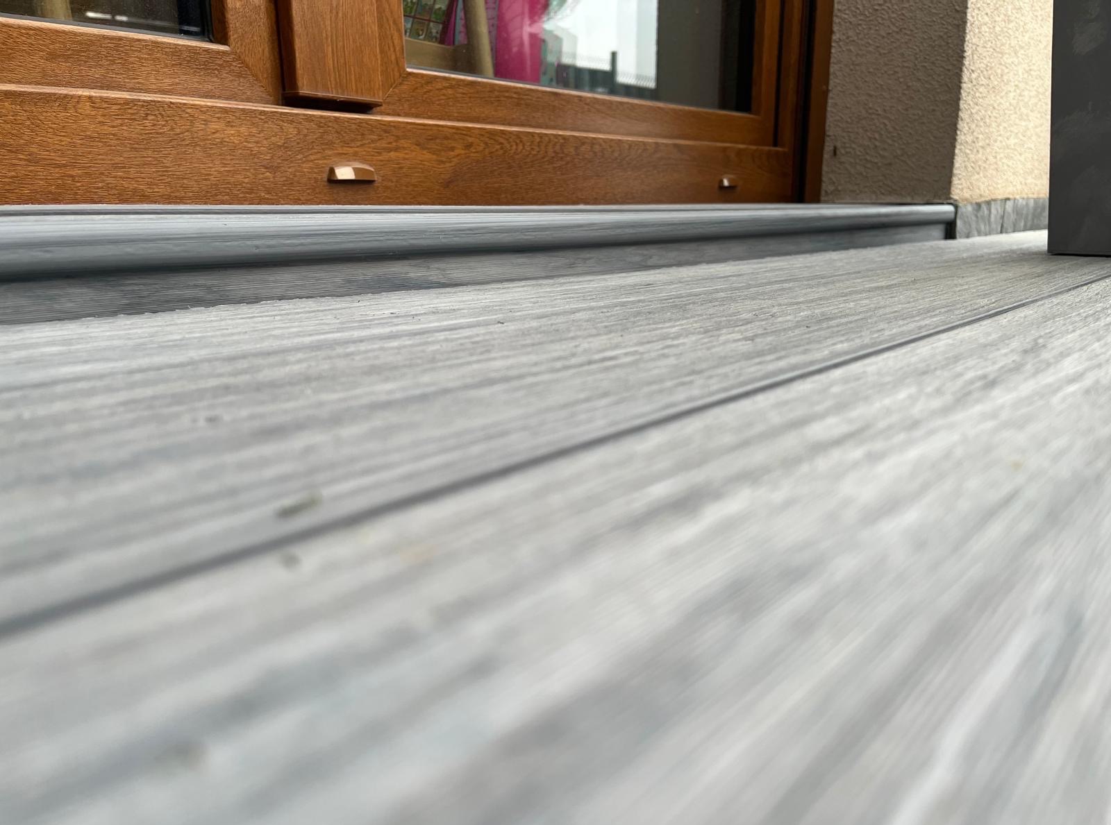 Szara deska kompozytowa tarasu przylegająca do brązowego okna z parapetem. Widok z niskiej perspektywy.