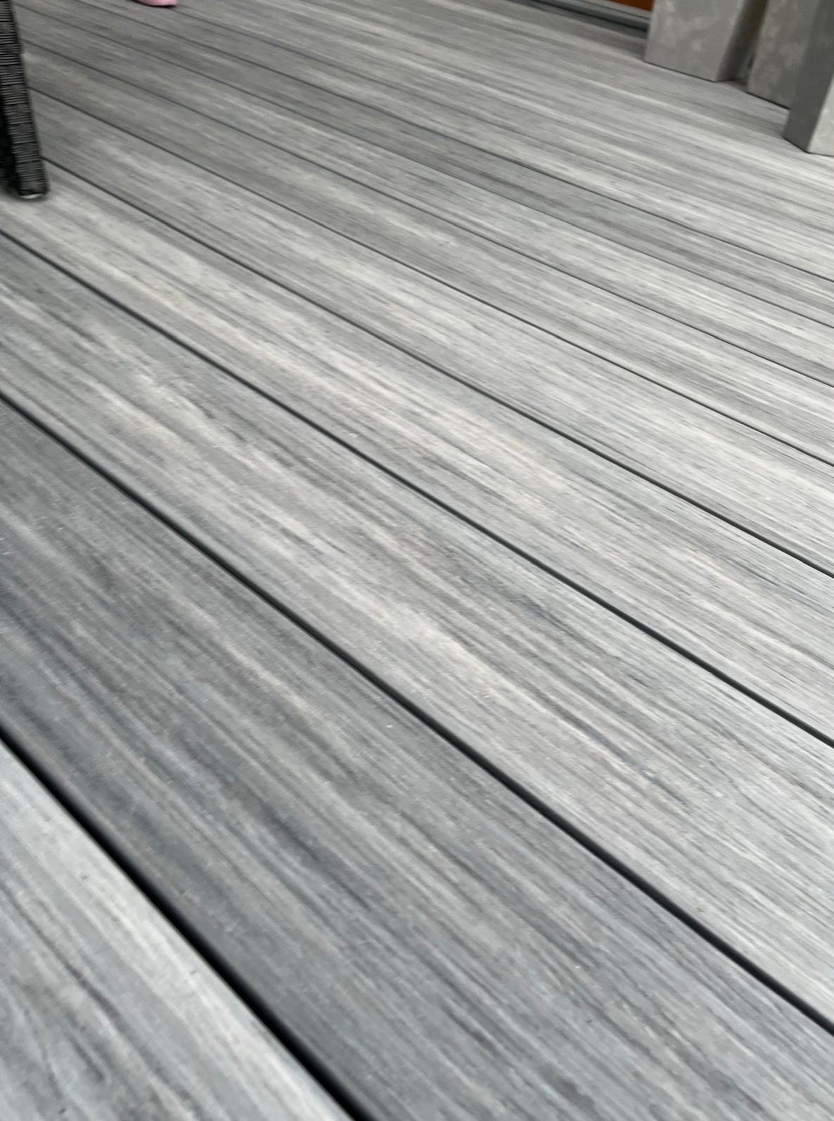 Szare deski tarasowe ułożone ukośnie, widoczne z bliska, z elementami mebli ogrodowych w tle. Delikatna faktura i subtelne cieniowanie podkreślają nowoczesny design.