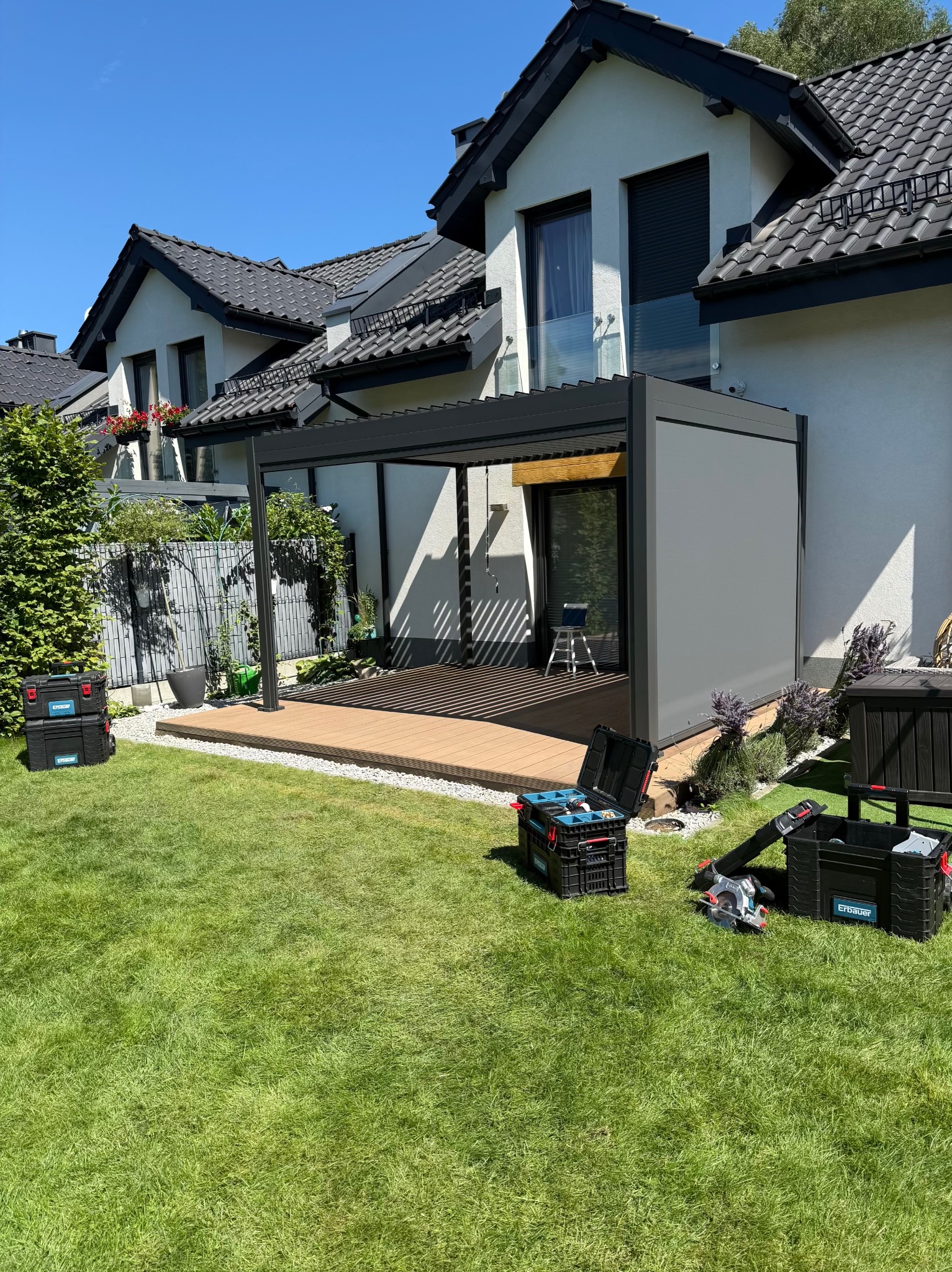 Nowoczesna pergola z szarymi panelami bocznymi i drewnianym tarasem. Na trawie skrzynki z narzędziami, sugerujące etap montażu. Dom w tle z czarnym dachem.