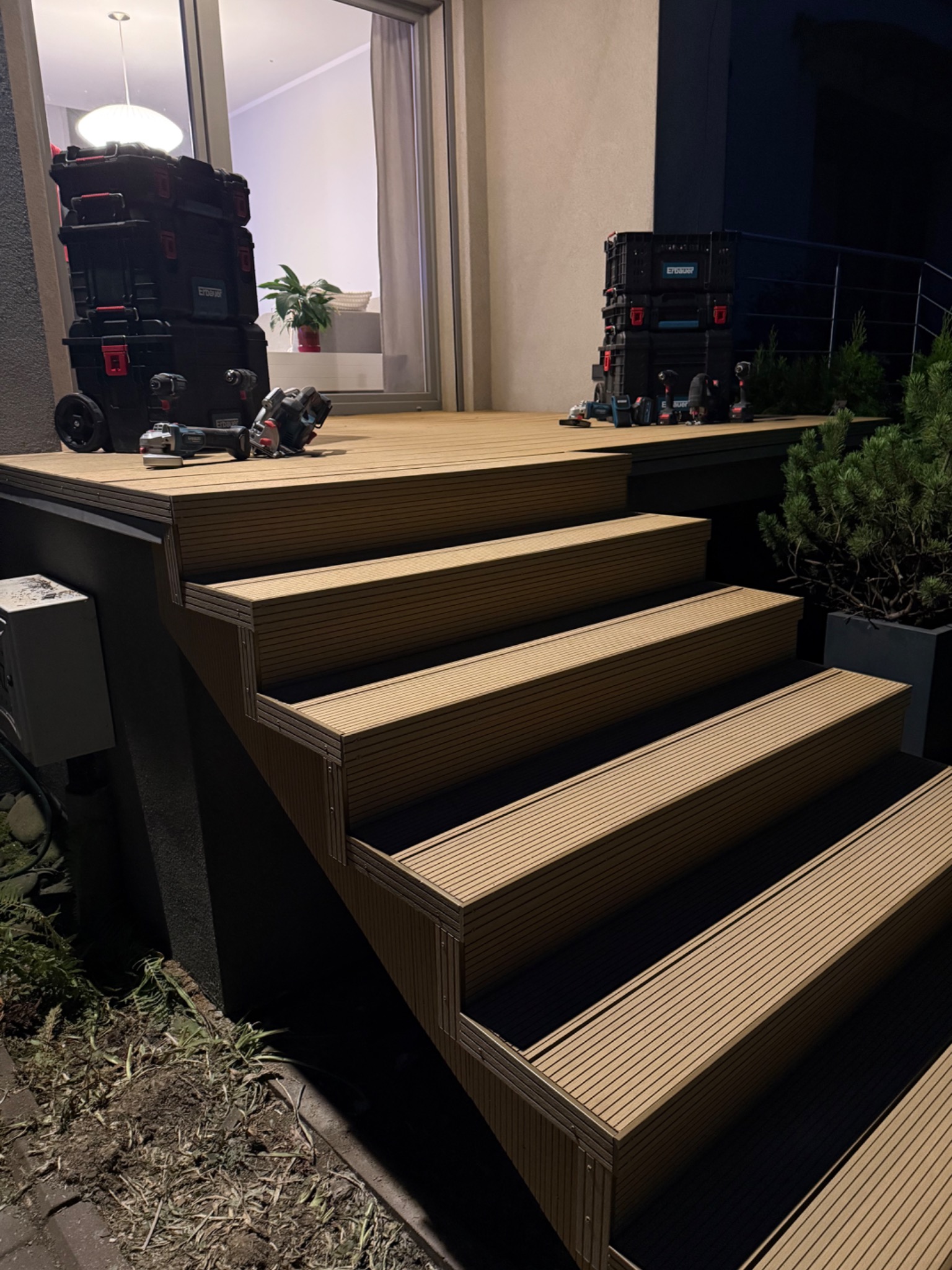 Schody z desek kompozytowych prowadzące na taras, na którym leżą skrzynki z narzędziami i elektronarzędzia. Widoczny fragment elewacji budynku i roślinność.