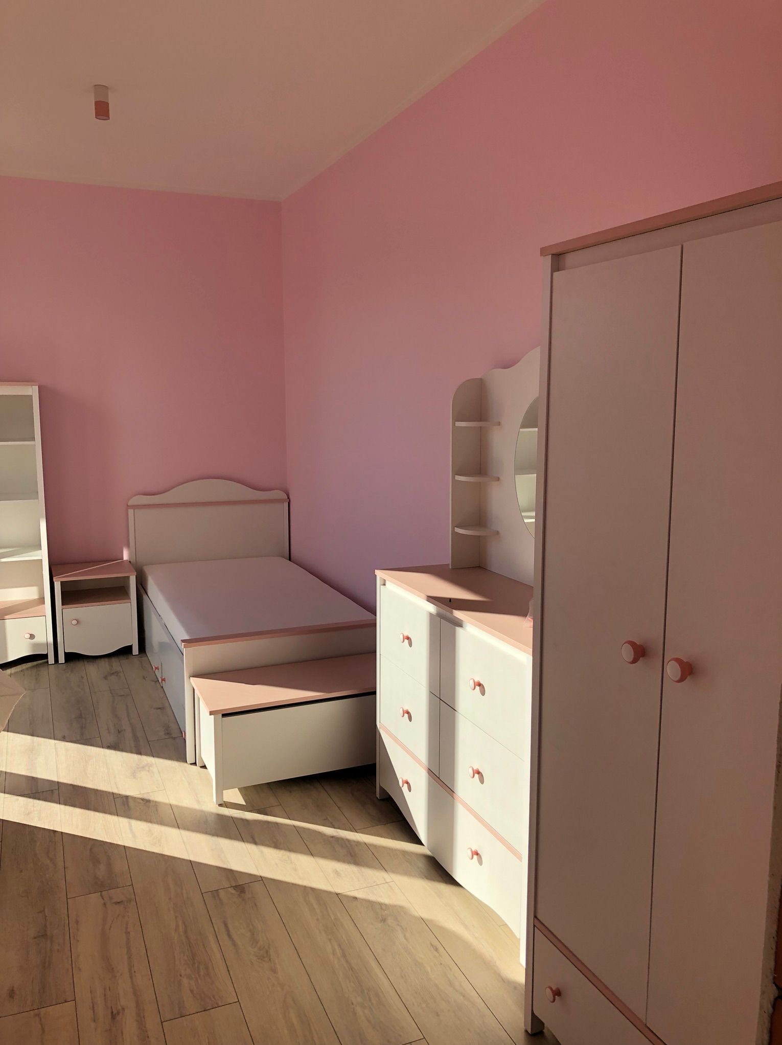 Pokój dziecięcy w pastelowych barwach: białe meble z różowymi akcentami, łóżko z pojemnikiem, komoda z lustrem i szafa, na drewnianej podłodze, ściany w odcieniach różu.