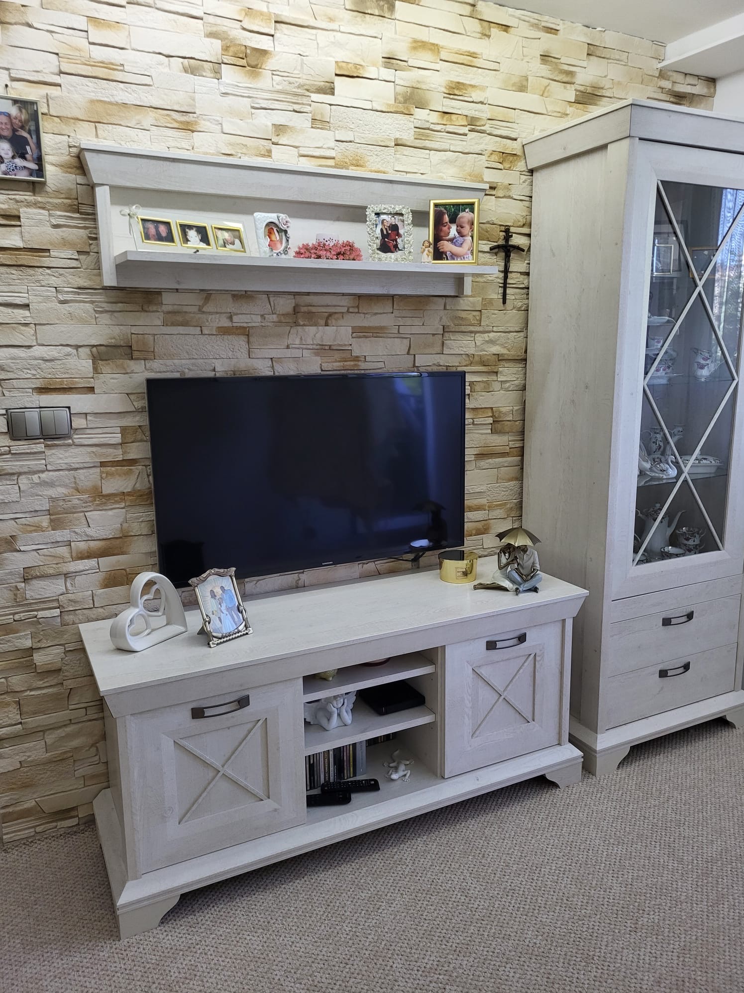 Jasny salon z meblami w stylu rustykalnym: szafka RTV z dekoracjami, witryna z przeszkleniami. Kamienna ściana z półką i ramkami.