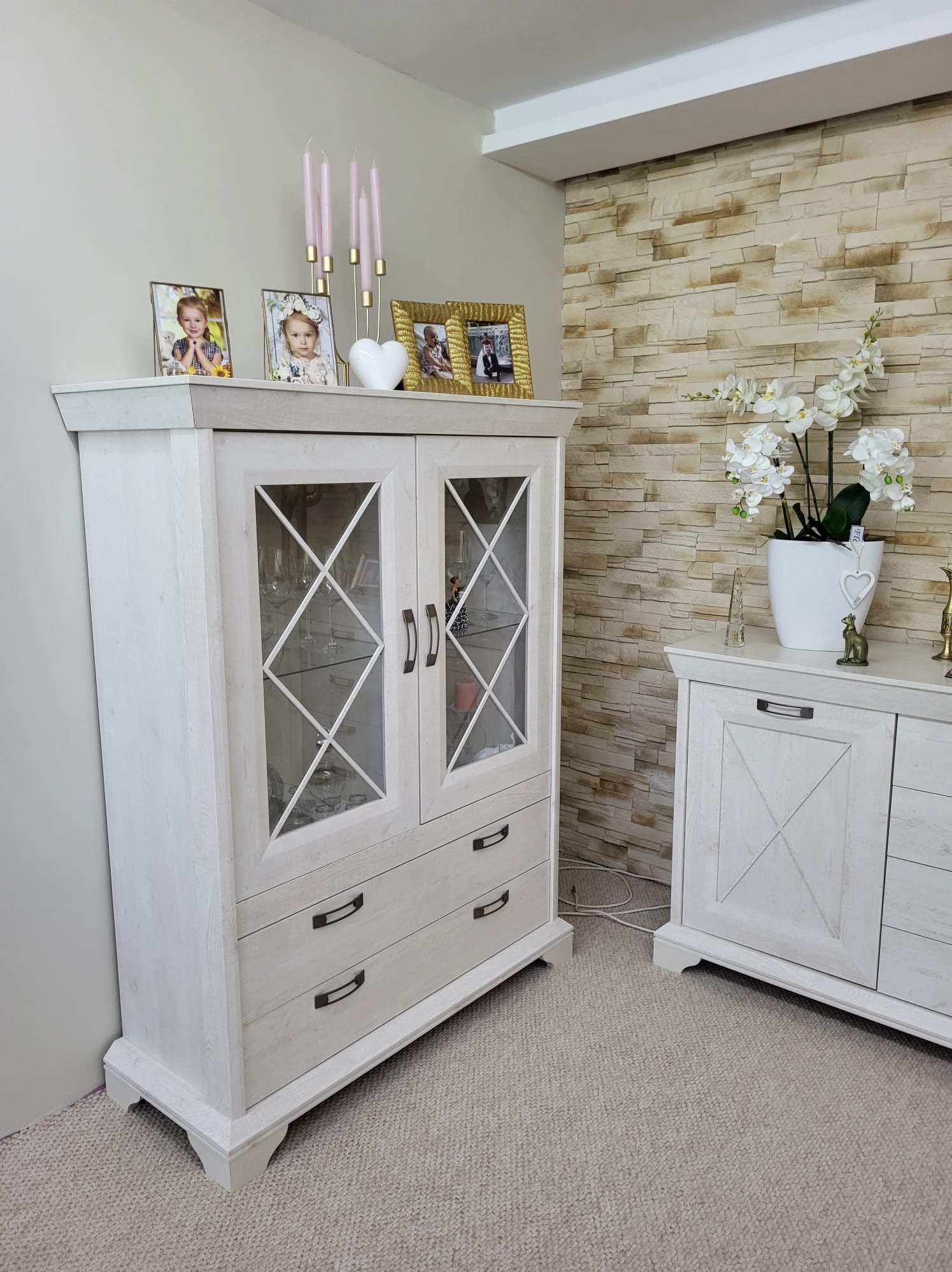 Biała, drewniana witryna z przeszkleniami i szufladami, ustawiona w pokoju z dekoracyjną ścianą z kamienia i komodą. Na witrynie zdjęcia i świeczniki.