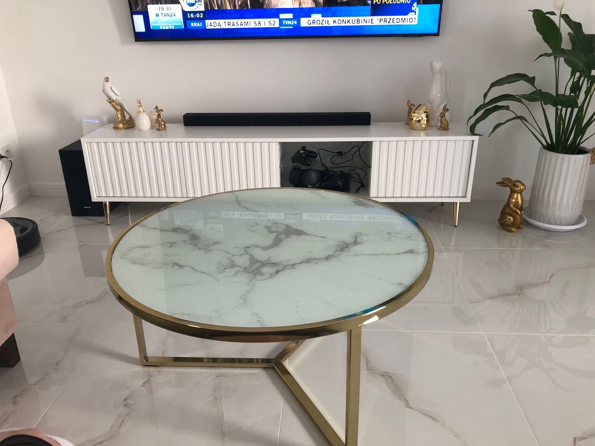 Stylowy salon z marmurową podłogą i okrągłym stolikiem kawowym ze złotymi nogami. W tle biała szafka RTV, telewizor i dekoracje.