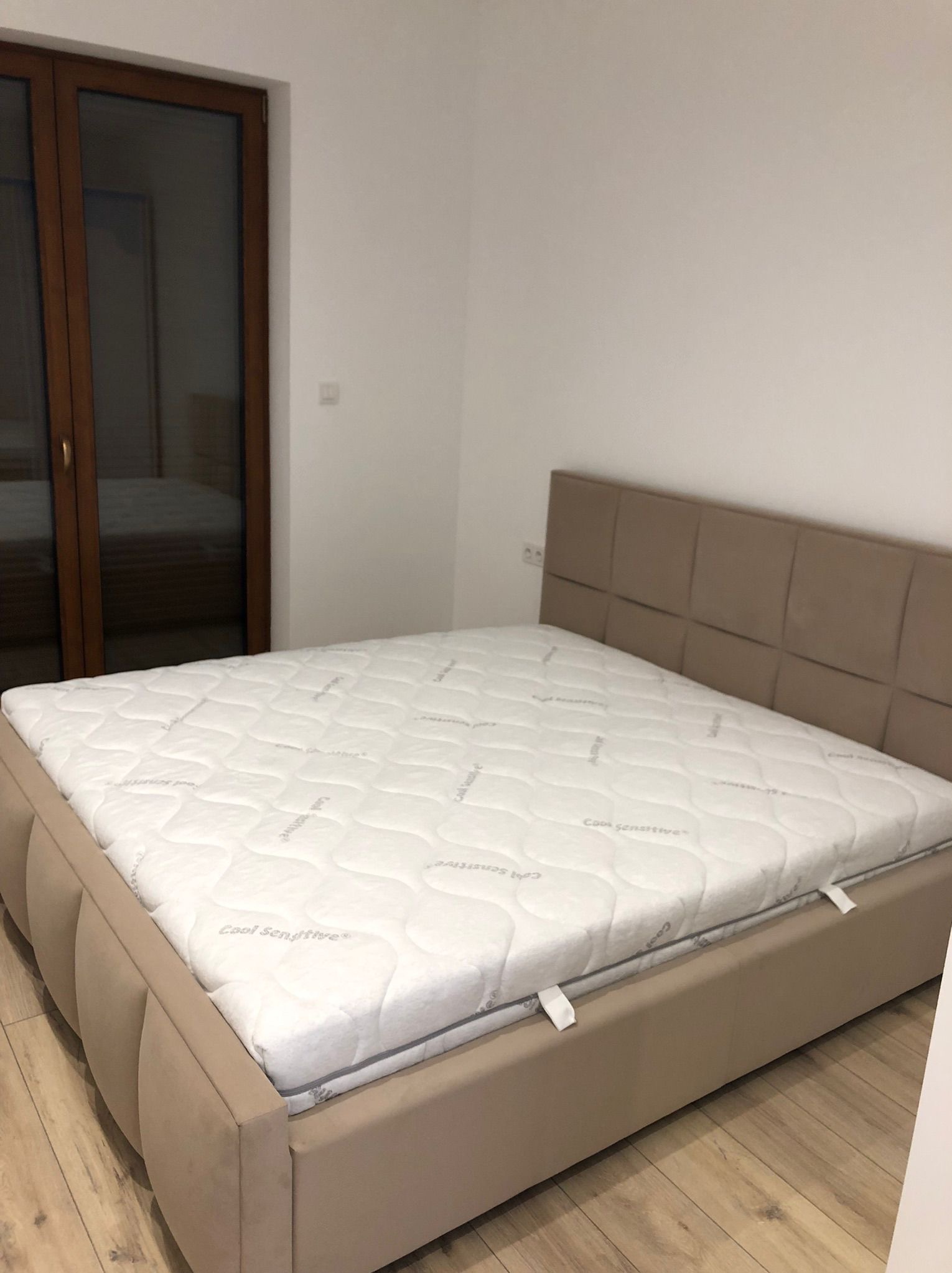 Beżowe łóżko z tapicerowanym zagłówkiem i materacem, widoczne na tle białej ściany i drewnianej podłogi. W tle okno balkonowe.