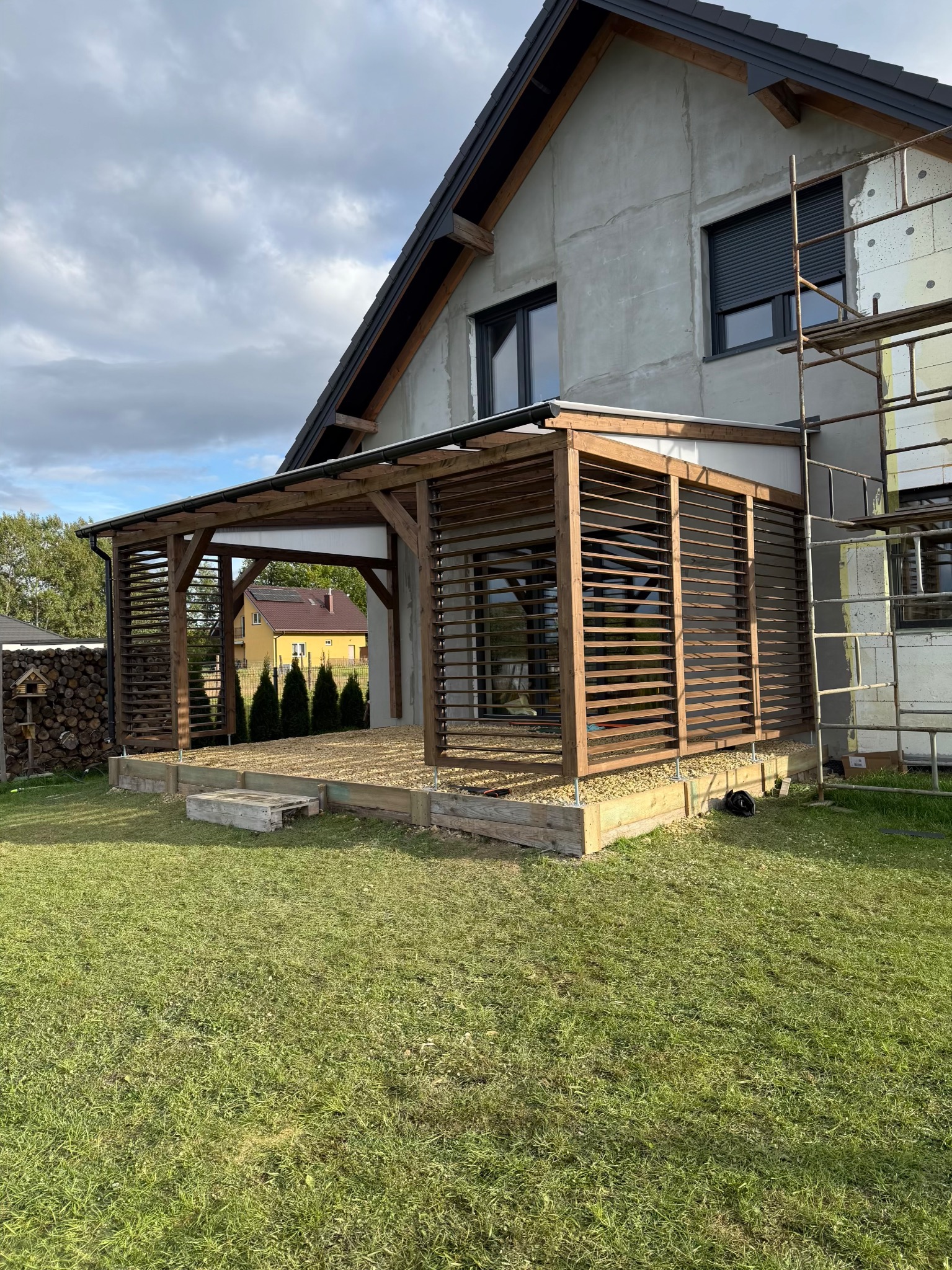 Drewniana pergola z lamelami, przylegająca do domu w stanie surowym. Widoczny podest z kamyczkami, trawnik i stos drewna. Częściowo widoczne rusztowanie.