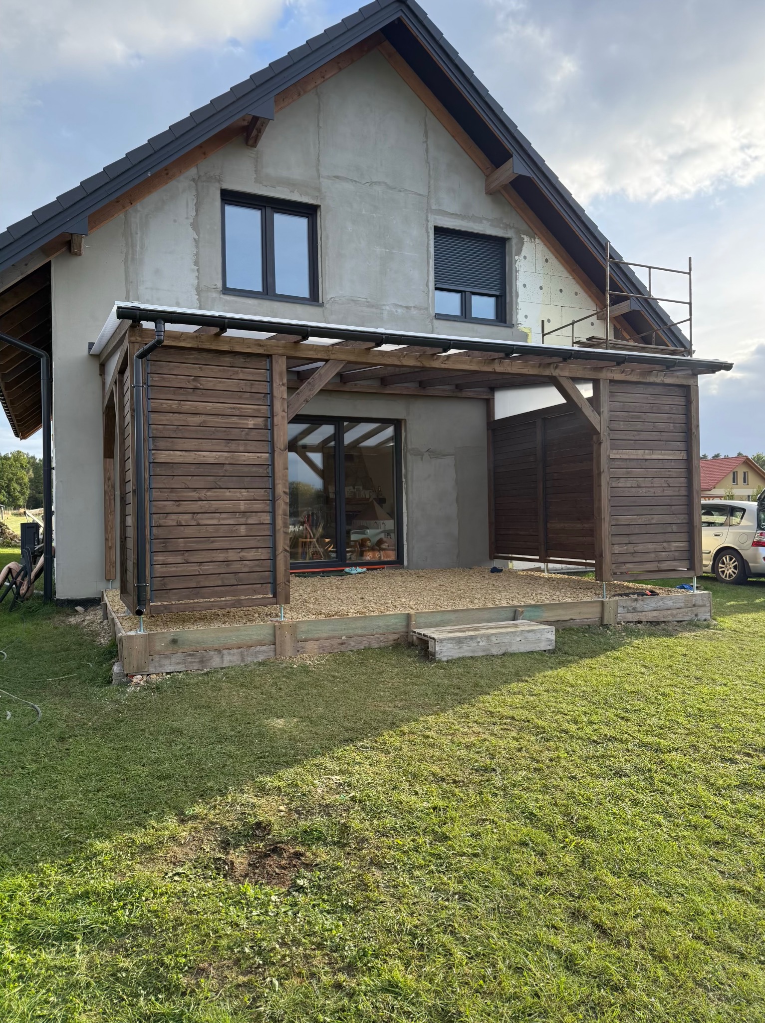 Drewniana pergola z zabudową częściową na tarasie przy domu w trakcie budowy, z widocznym ociepleniem na elewacji i rusztowaniem. Częściowo wysypany żwir na tarasie.