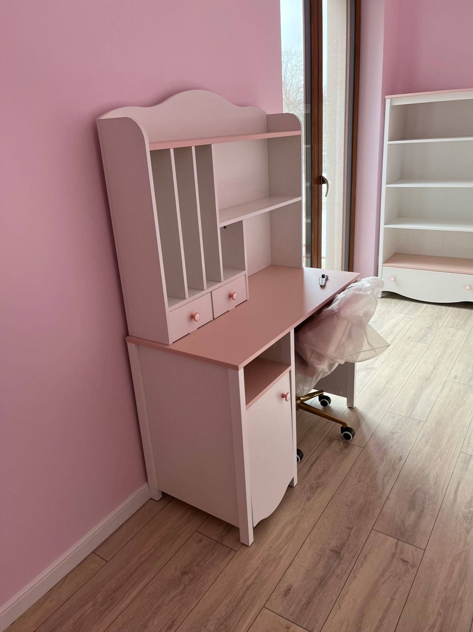 Różowo-białe biurko z nadstawką i krzesłem na kółkach, ustawione w pokoju z różowymi ścianami i drewnianą podłogą. W tle biały regał.