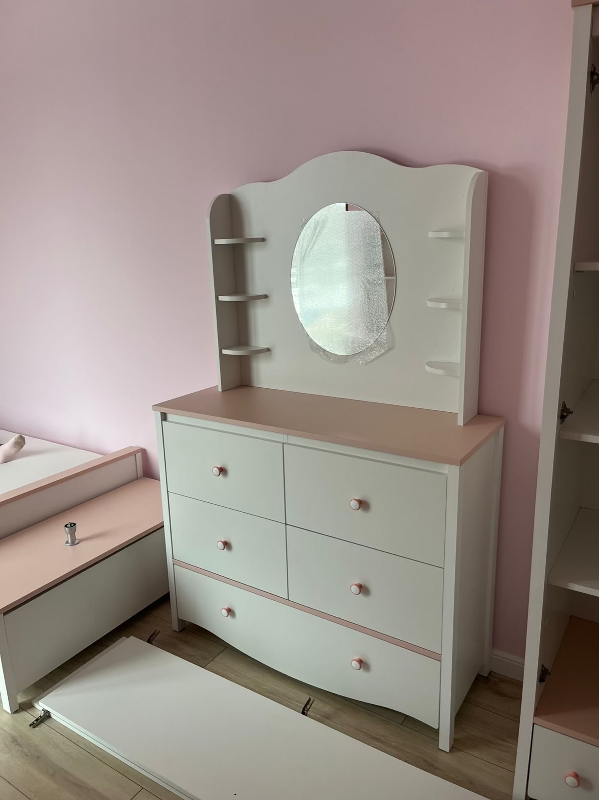 Biała komoda z owalnym lustrem i półkami, elementy łóżka dziecięcego w pokoju z różowymi ścianami. Meble w trakcie montażu, widoczne niedokończone elementy.