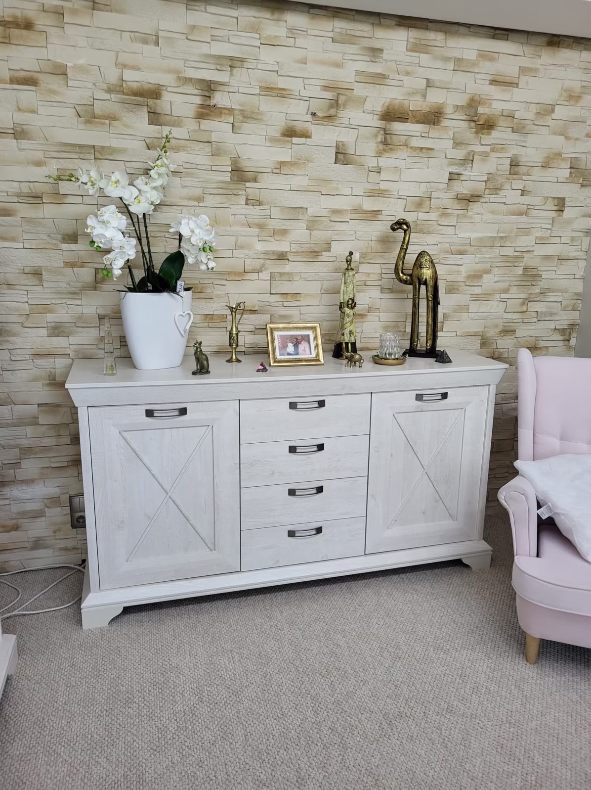 Biała komoda z dekoracjami (figurki, kwiaty) na tle kamiennej ściany i różowego fotela. Mebel z frezowanymi drzwiami i metalowymi uchwytami.