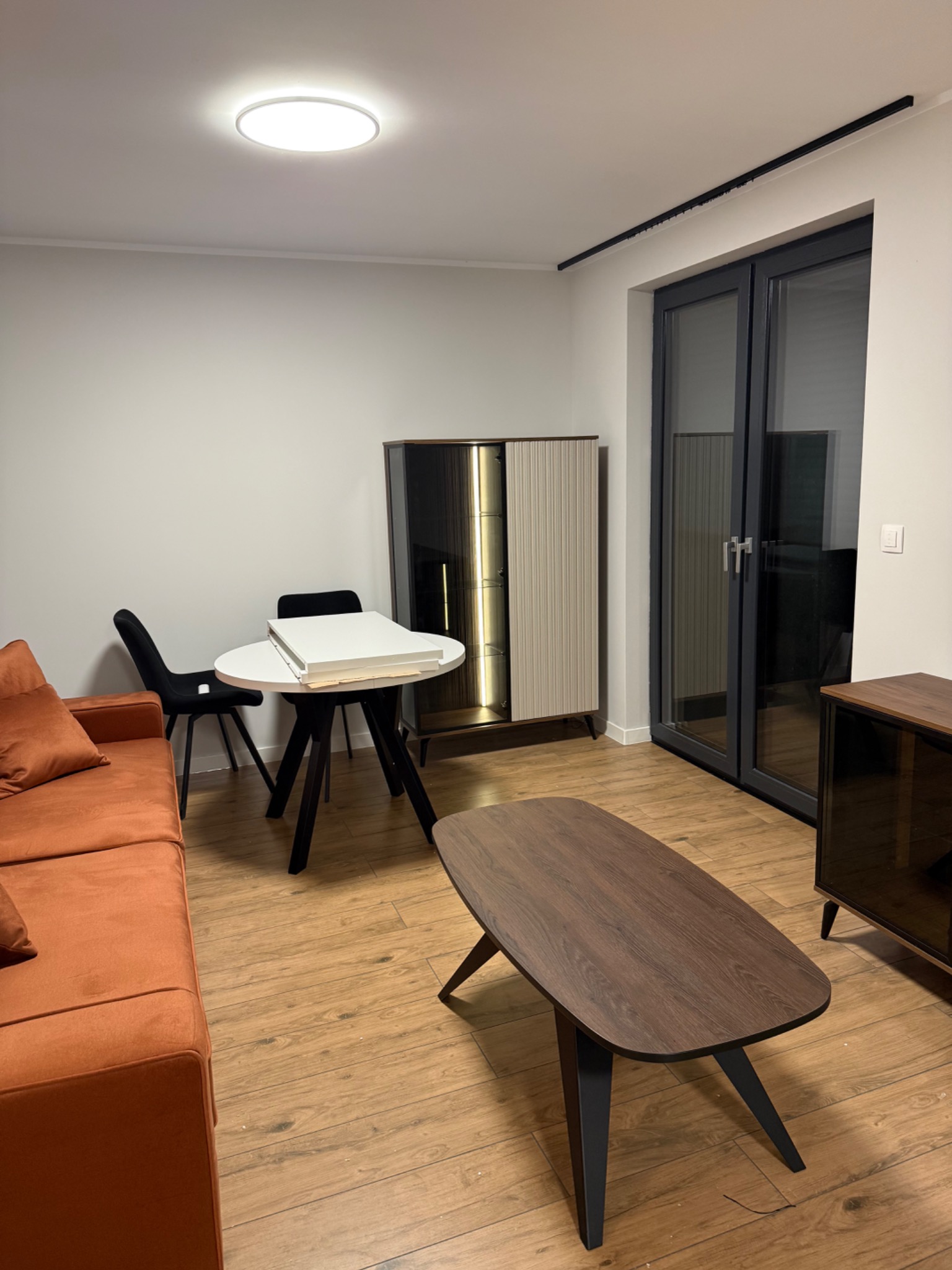 Jasne wnętrze z pomarańczową sofą, stolikiem kawowym i szafką. Na okrągłym stole leżą elementy meblowe, prawdopodobnie do montażu. Widoczne drzwi balkonowe.