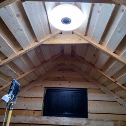 Rafał Marciniak - Montaż okrągłej lampy sufitowej w drewnianym wnętrzu. Widoczny fragment okna i narzędzie na wysięgniku. Ujęcie od dołu.