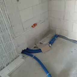 Surowy stan: kable elektryczne w białych rurkach na ścianie z pustak&oacute;w, niebieskie rury w podłodze, pomarańczowe puszki elektryczne. Instalacja w trakcie prac.