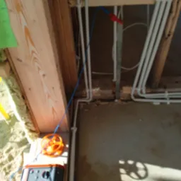 Pomiary instalacji elektrycznej w surowym wnętrzu. Widoczne przewody, miernik i elementy konstrukcji drewnianej. Niebieski przewód pomiarowy.