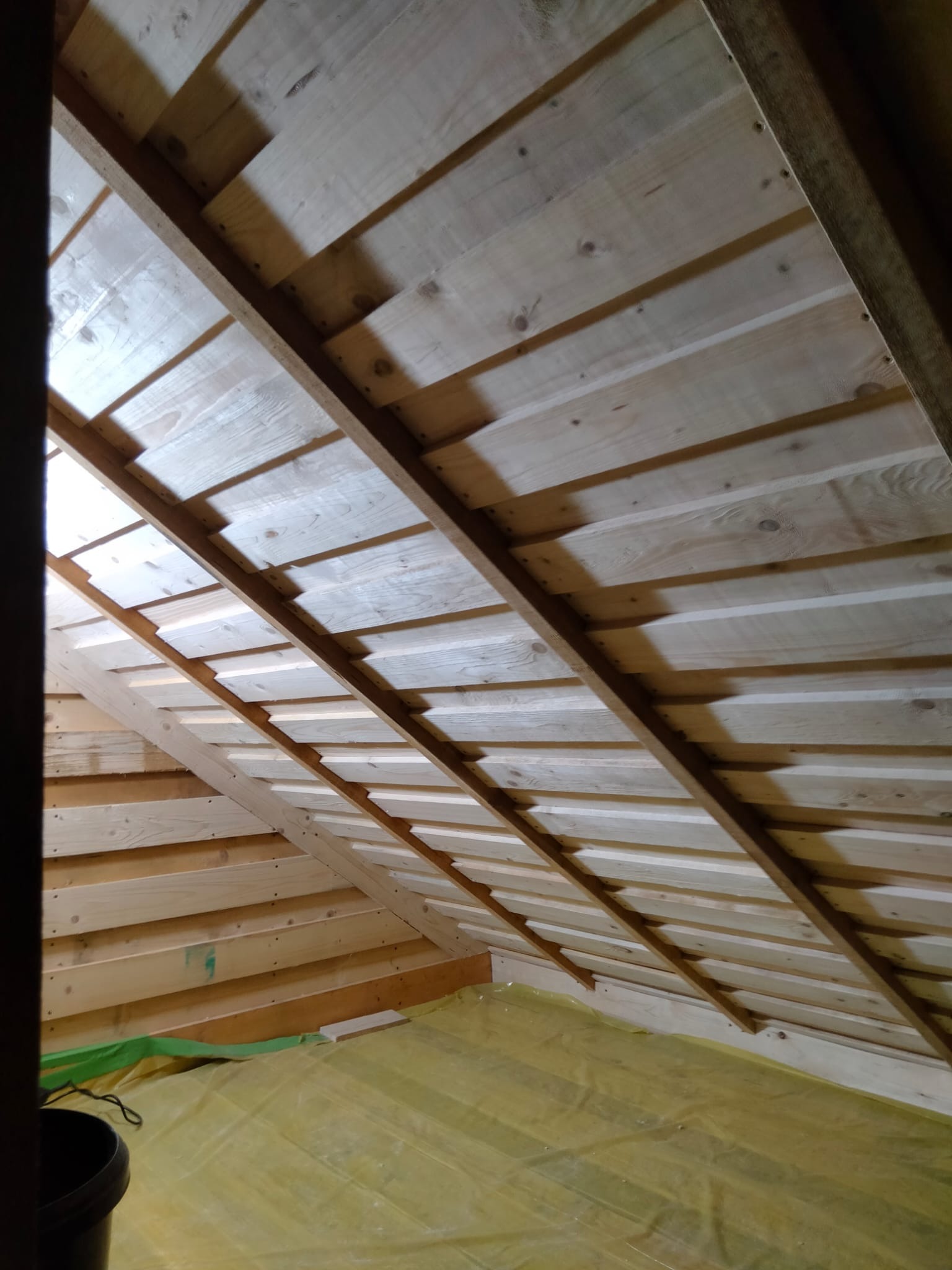 Drewniana konstrukcja dachu z widocznymi elementami więźby i deskowaniem, zabezpieczona folią budowlaną na podłodze. Układ belek tworzy regularny wzór.