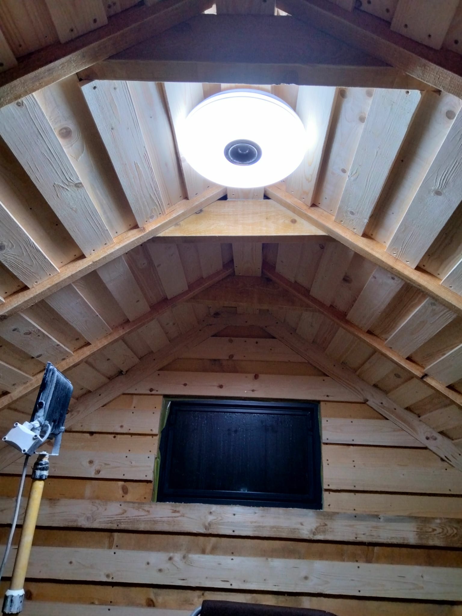 Montaż okrągłej lampy sufitowej w drewnianym wnętrzu. Widoczny fragment okna i narzędzie na wysięgniku. Ujęcie od dołu.