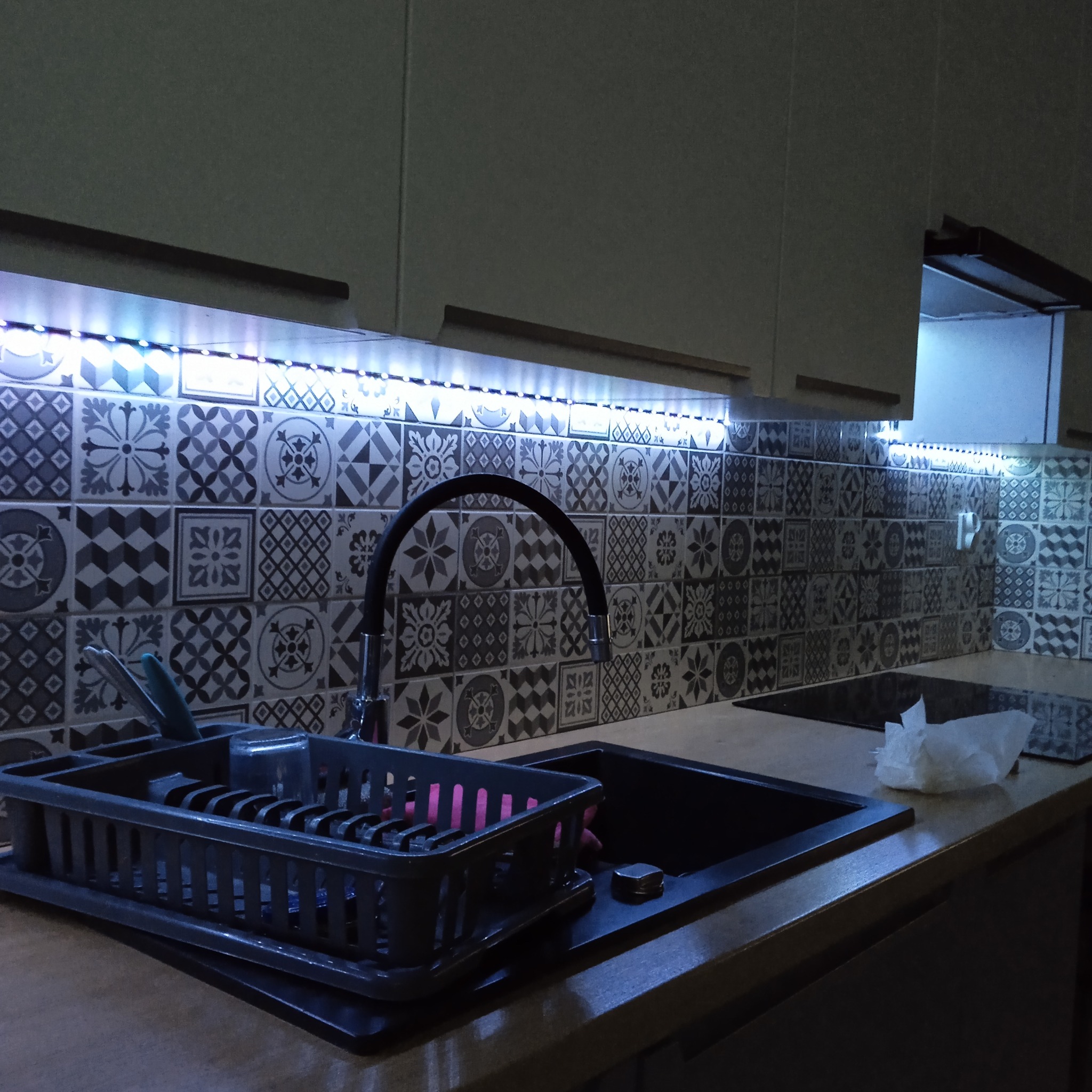 Kuchnia po montażu taśmy LED pod szafkami, oświetlającej wzorzyste płytki nad blatem i czarny zlew z ociekaczem. Nowoczesne rozwiązanie oświetleniowe.