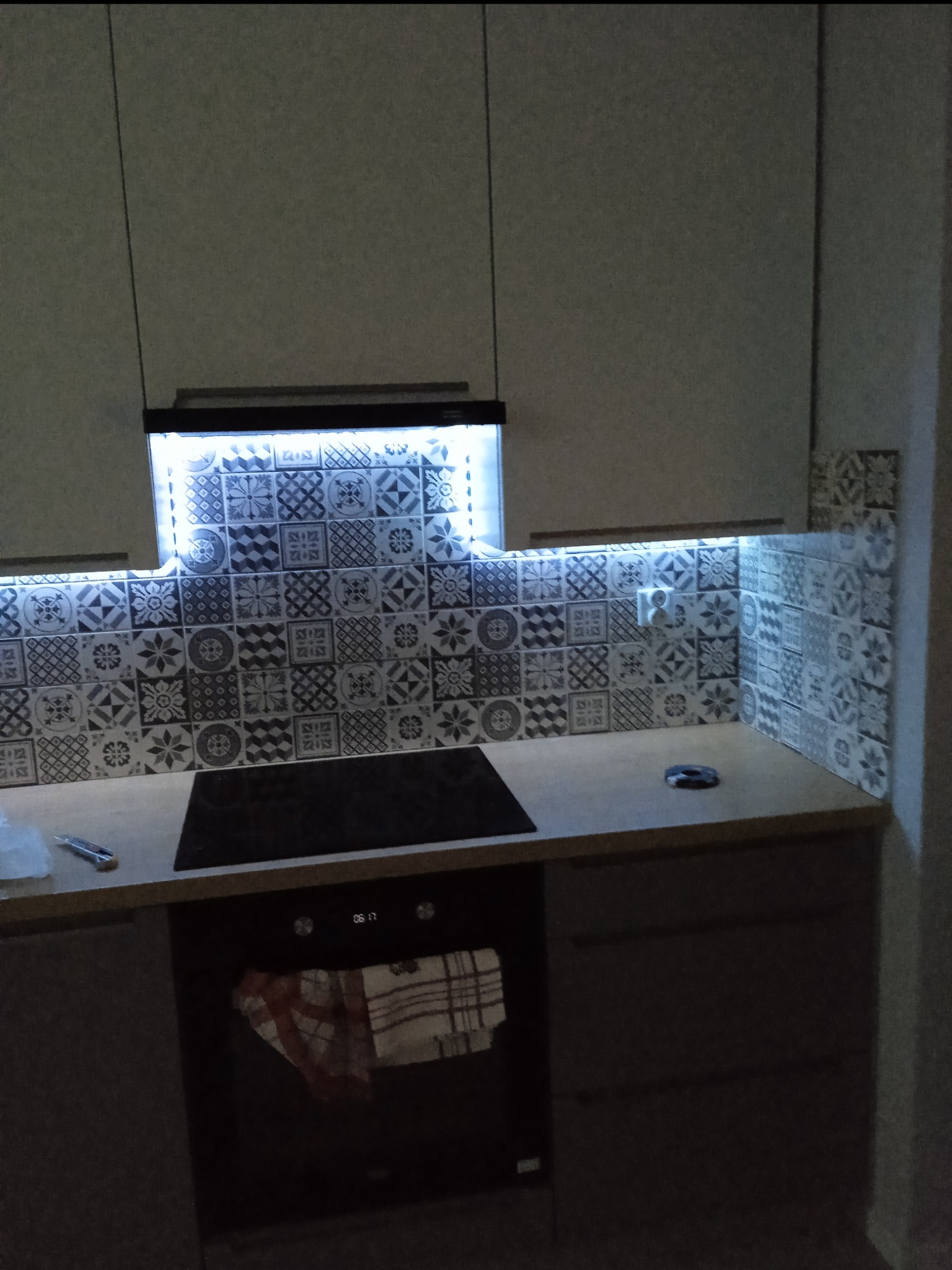 Nowoczesna kuchnia z podświetleniem LED nad blatem i płytą indukcyjną. Dekoracyjne płytki nadają wnętrzu unikalny charakter. Widoczny fragment piekarnika.